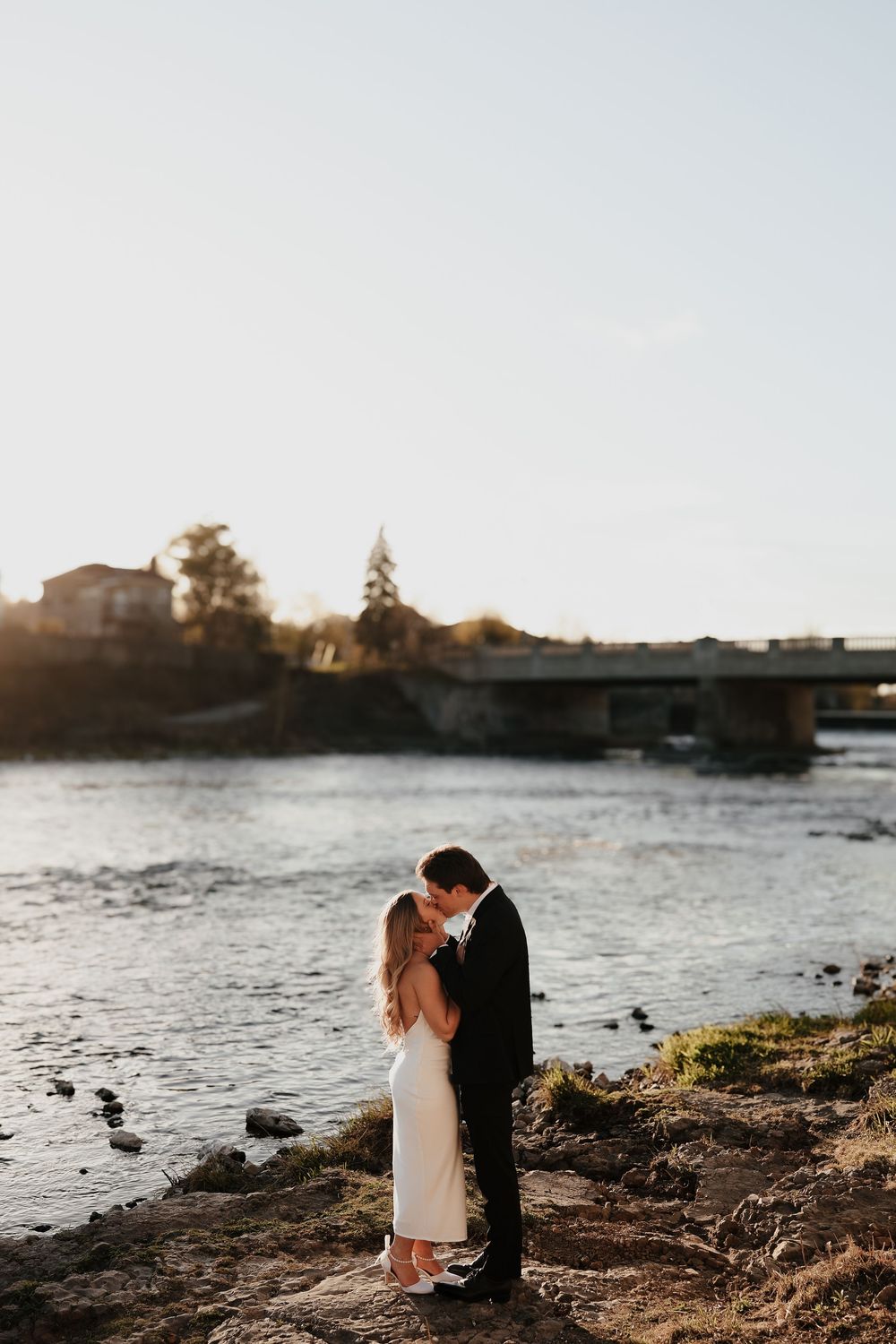 A romantic sunset wedding couple embraces on a rocky shoreline near a tranquil lake.