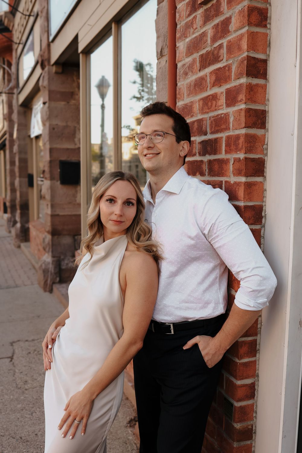 A couple poses playfully against a red brick wall and white columns in coordinated white outfits.