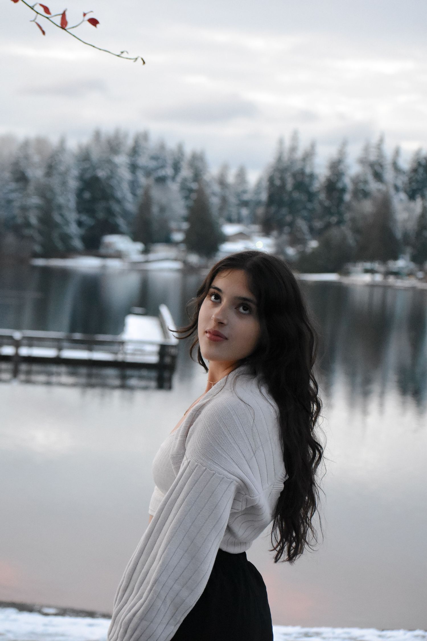 Person posing against a serene winter lake backdrop with snow-covered trees in the background.