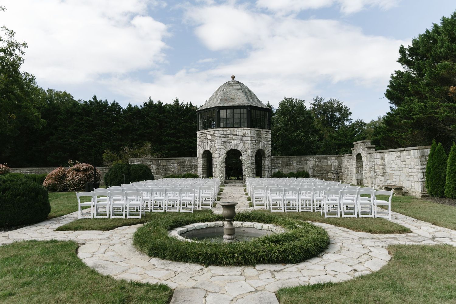 A panoramic view of a stone gazebo and white chairs arranged in a semi-circle for an outdoor wedding ceremony.