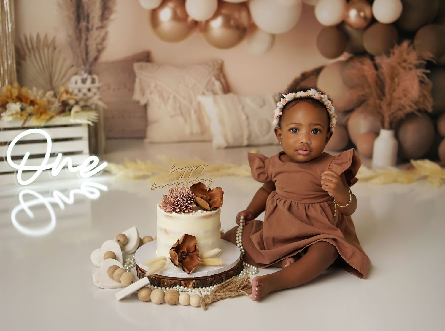 Baby girl in a brown ruffled dress and floral headband sitting beside a cake at a boho first birthday cake smash photoshoot.