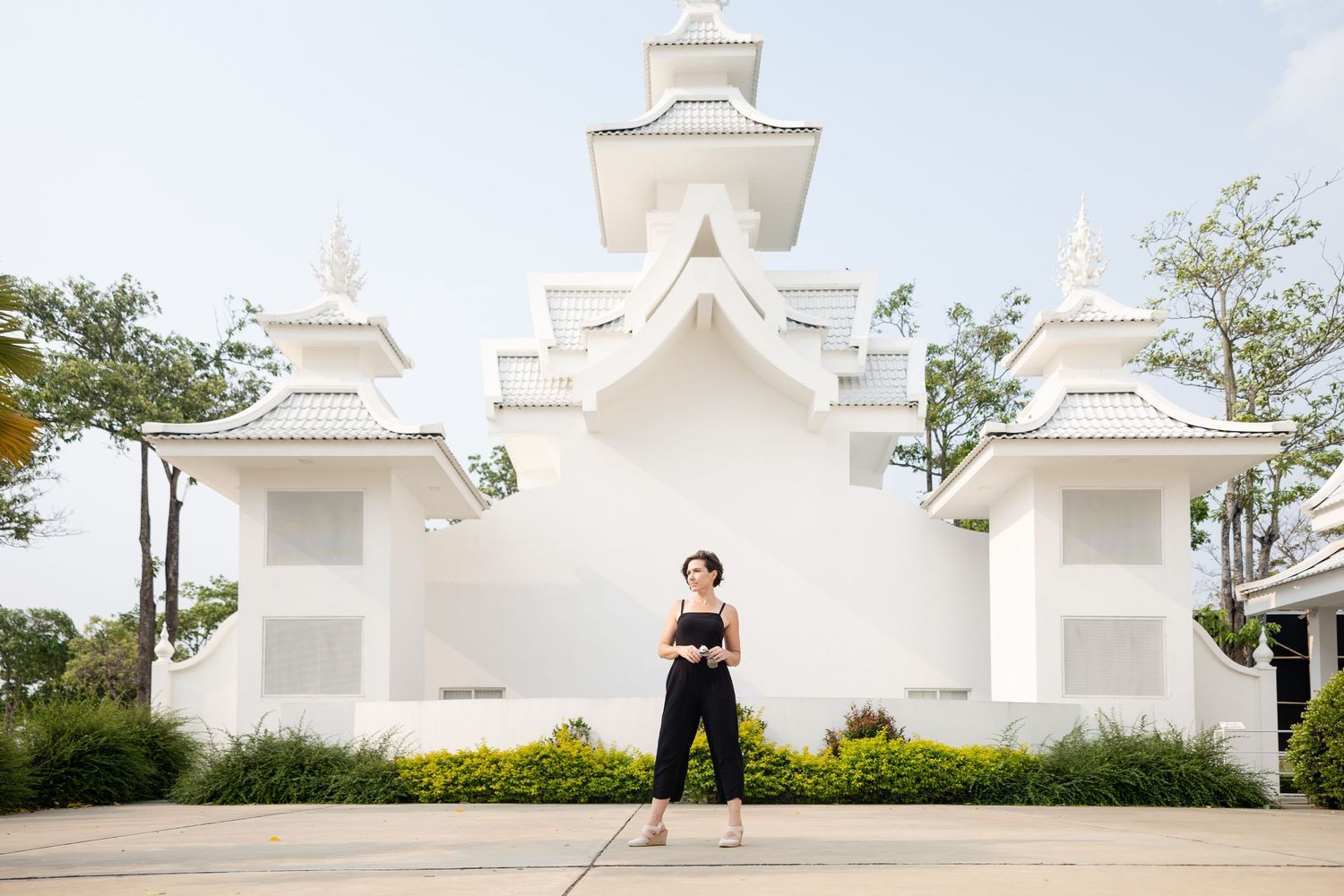 A person in black attire poses in front of a white architectural wall with ornate details in an outdoor garden setting.