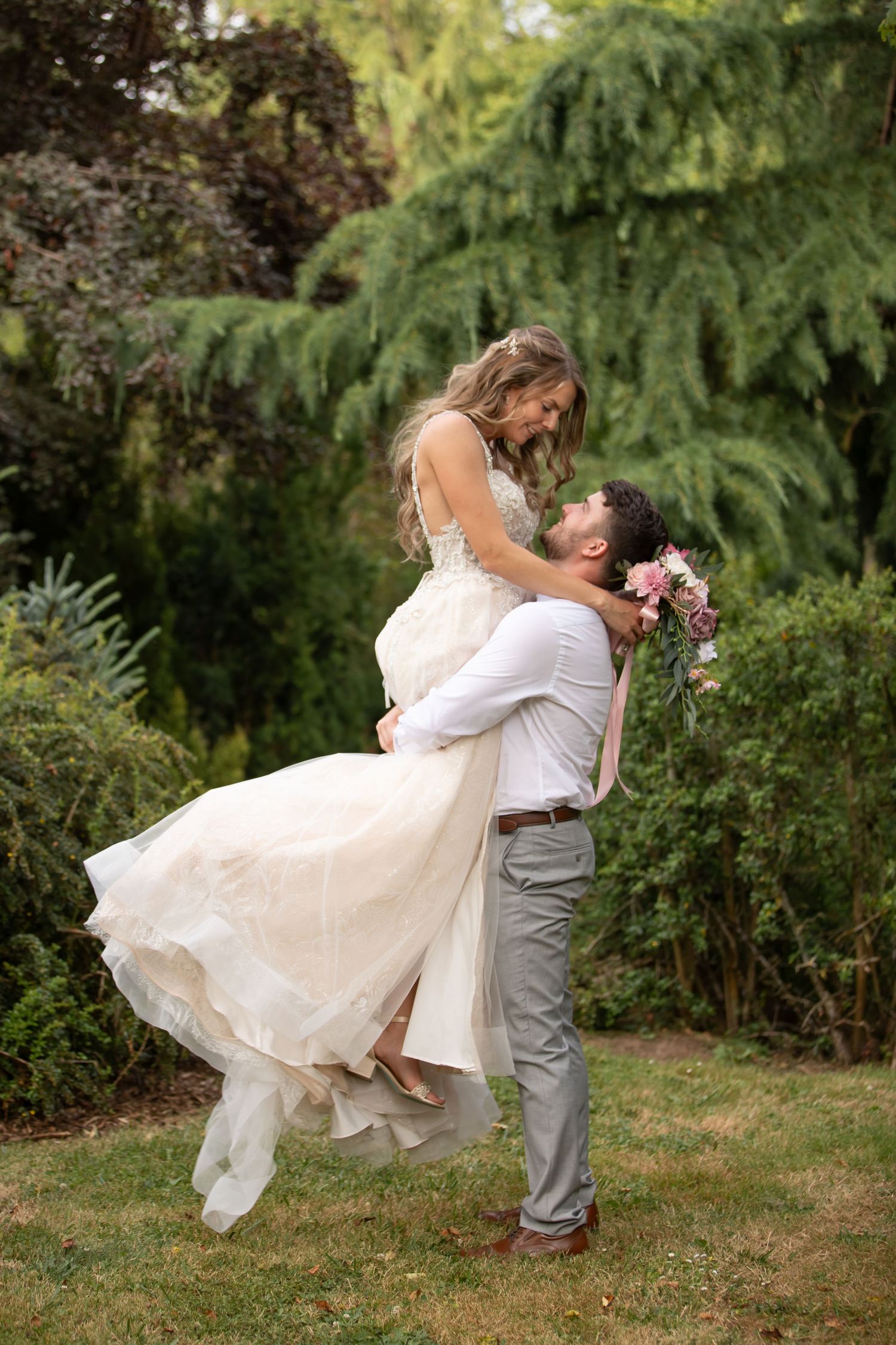 Romantic wedding couple embraces in a garden as groom lifts bride in white dress.