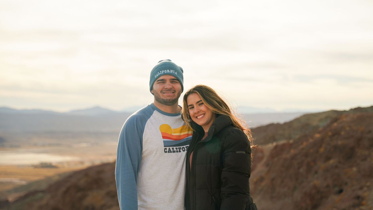 Couple bundled in winter clothes smile while standing on mountain vista.