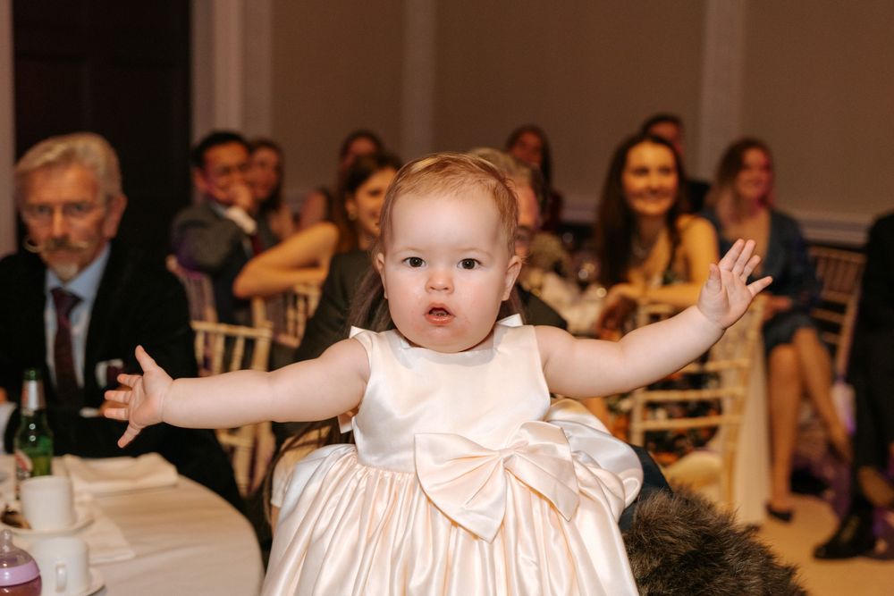 Adorable baby in a white dress with bow excitedly gestures during a wedding reception.