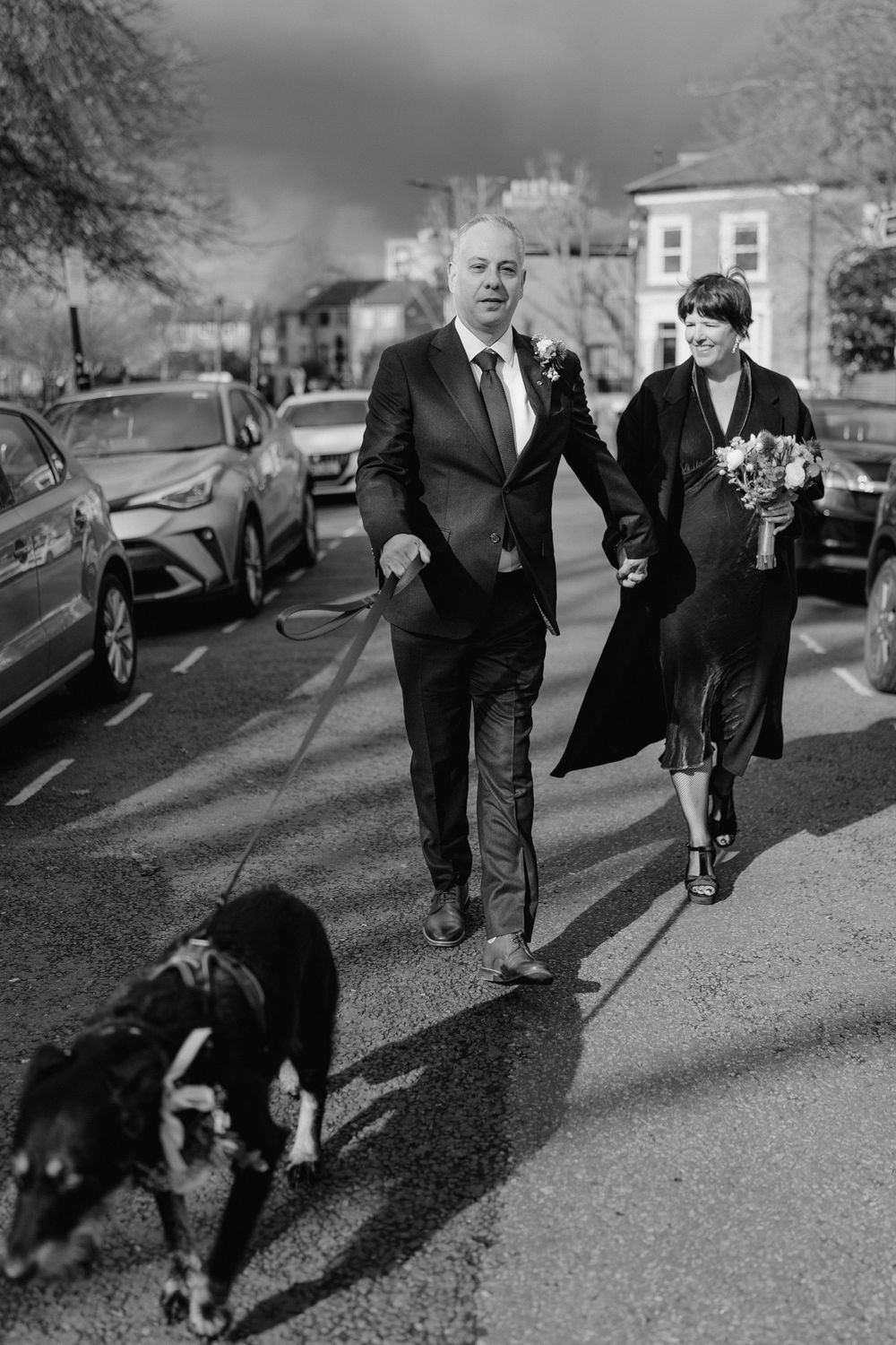 A couple walks down a street with a dog on a cloudy day.