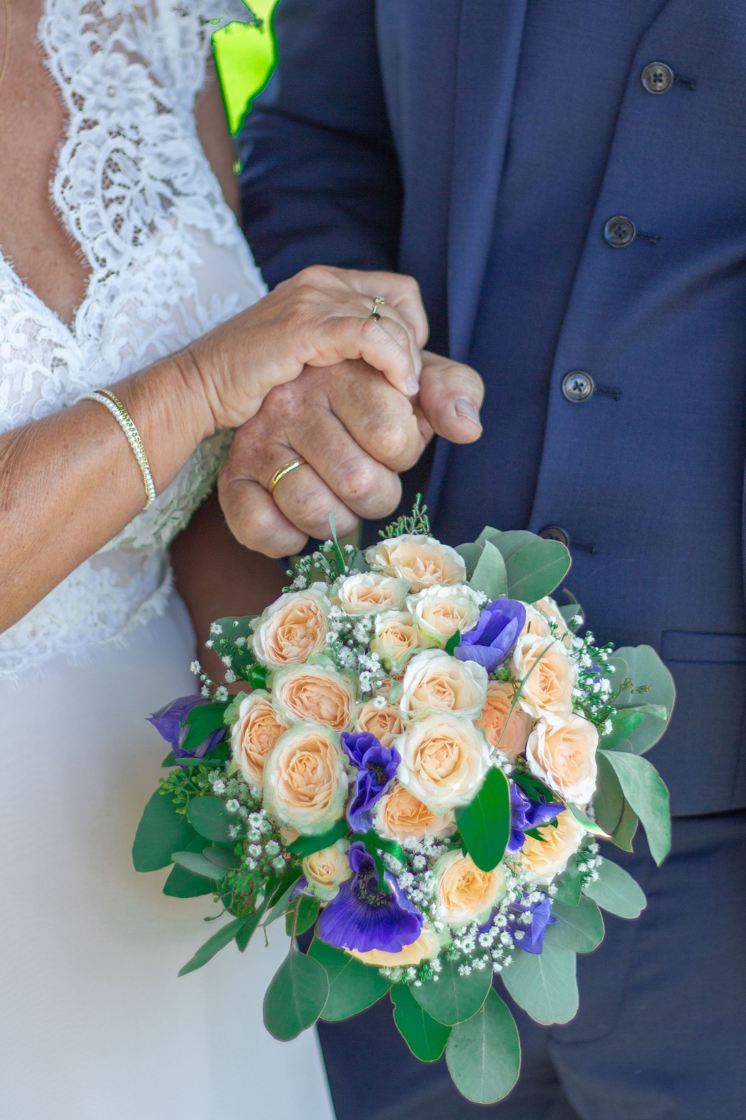 Wedding bouquet of peach roses and purple flowers held by couple's clasped hands.
