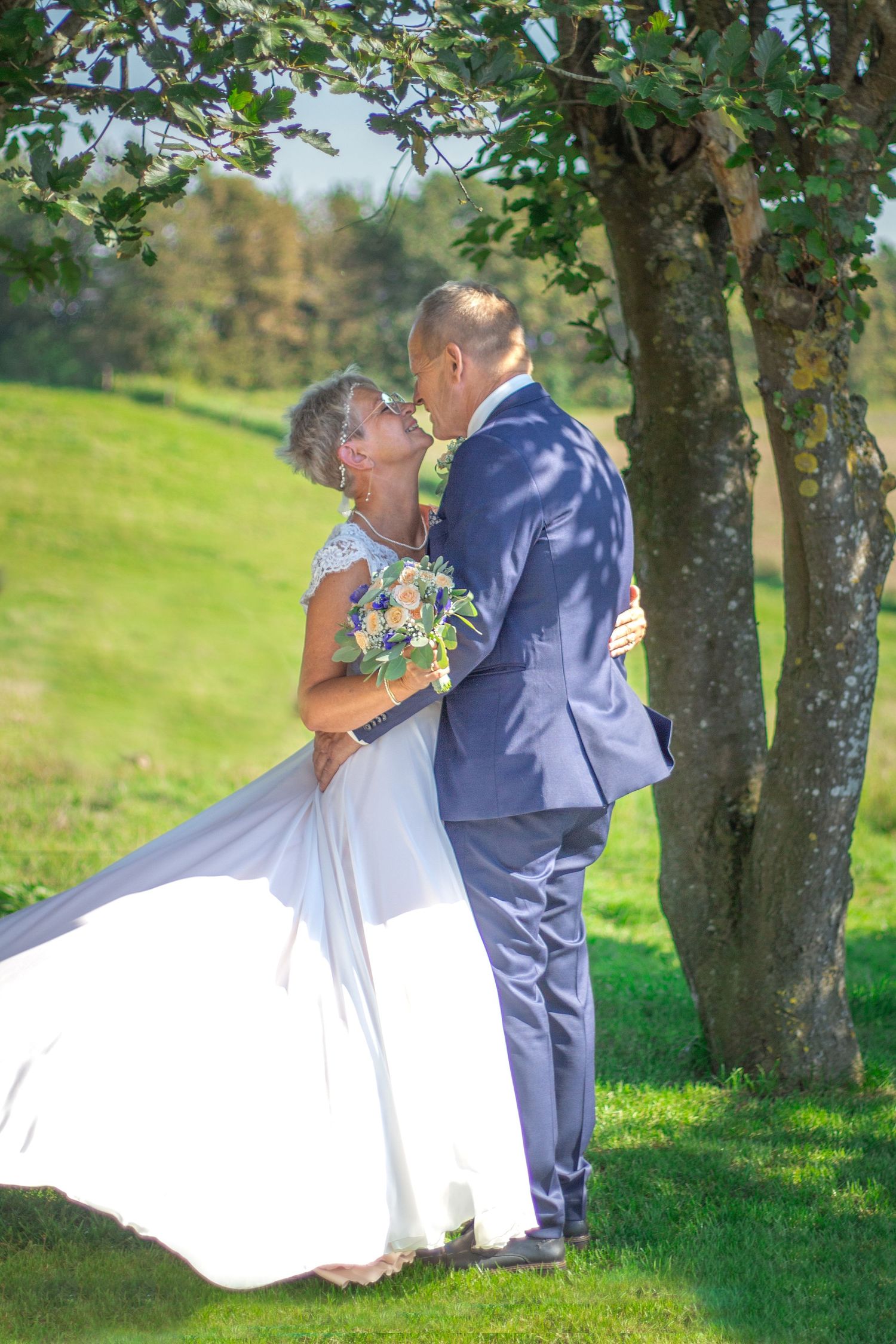Elegant couple in wedding attire sharing romantic moment under tree in sunny field.