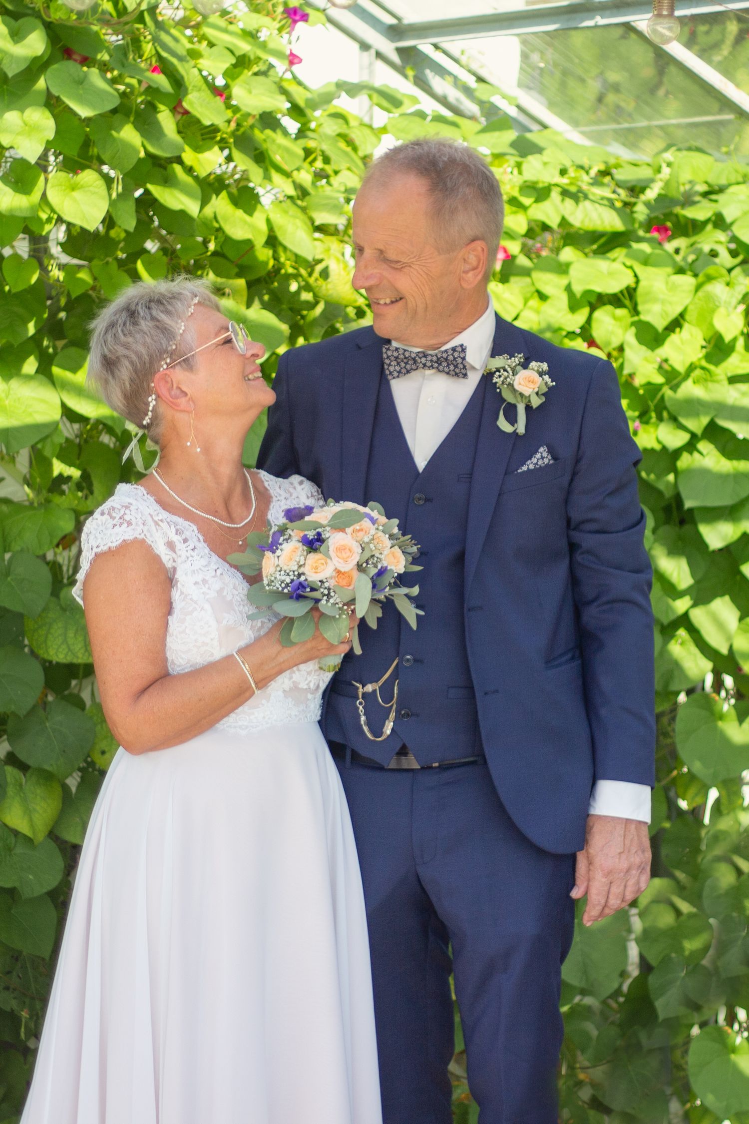 A couple in wedding attire share a joyful moment in front of lush greenery, with white dress and navy suit.