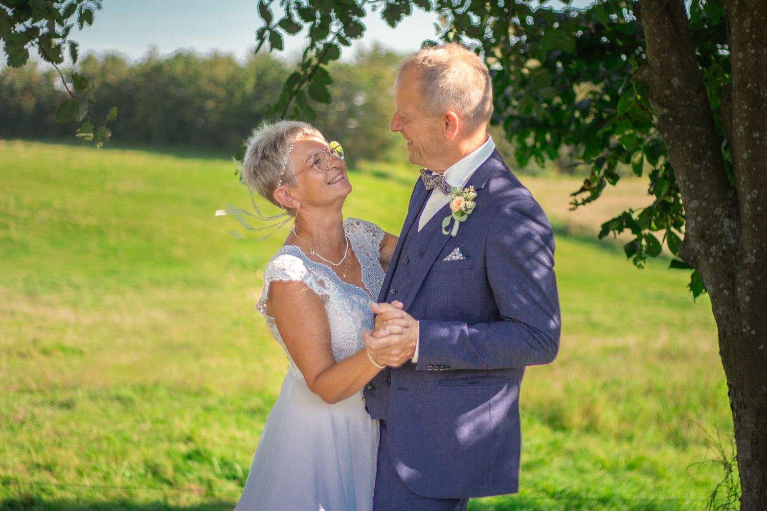 Mature couple dancing together in elegant wedding attire under sunlit tree.