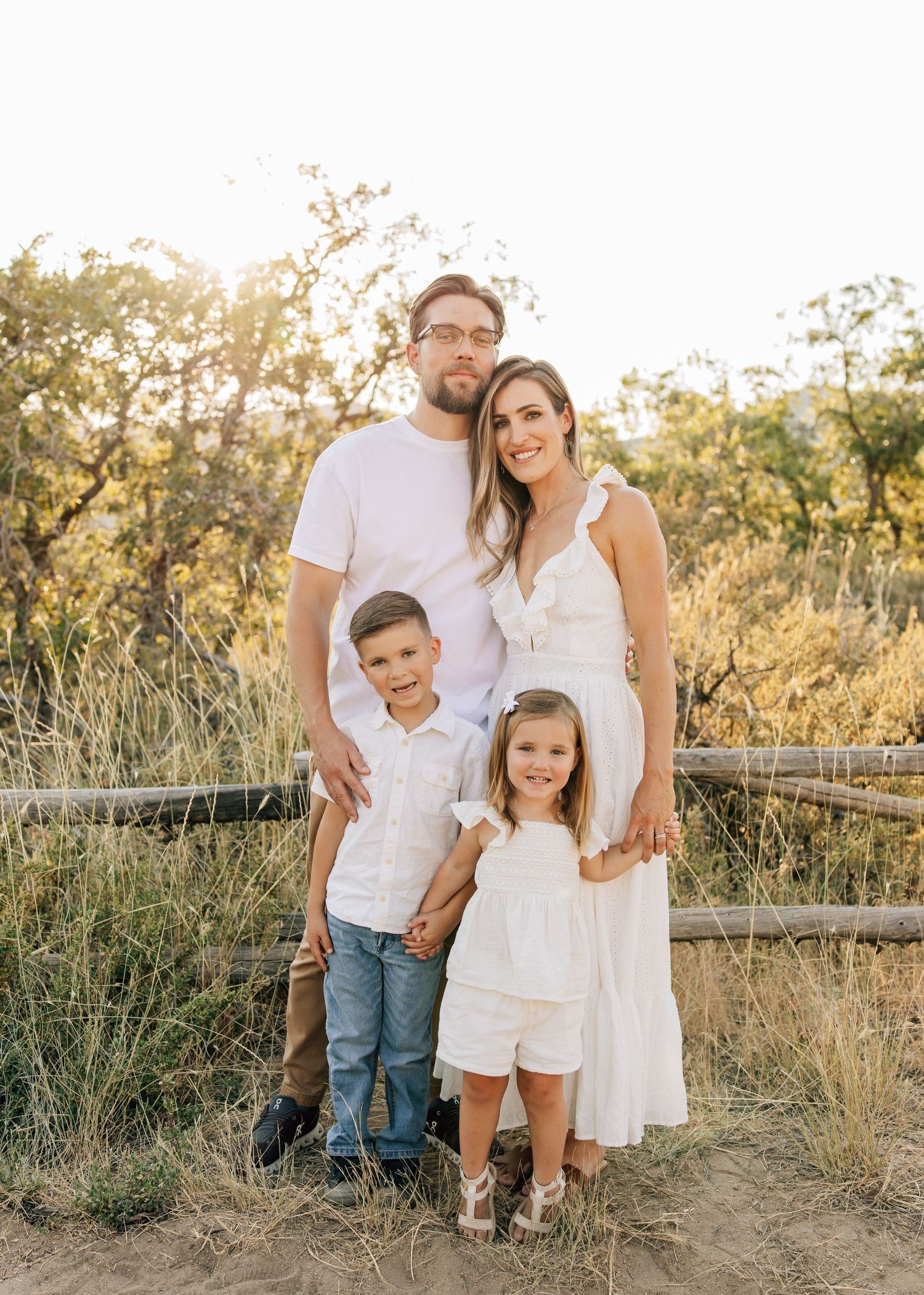 A family poses together in front of a rustic wooden fence at golden hour with tall grass and sunlit trees in the background.