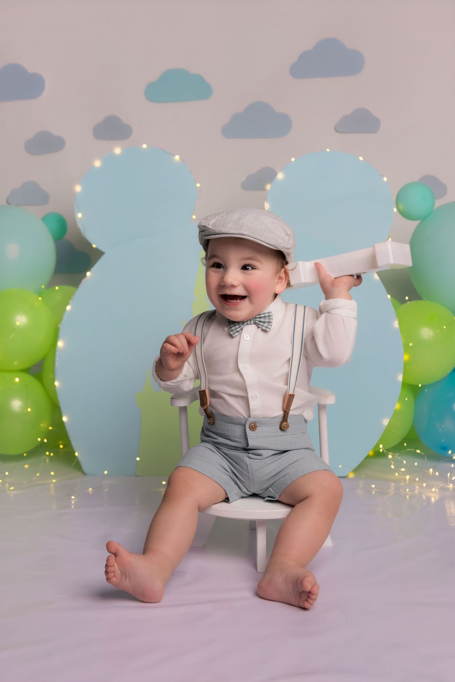 Toddler in vintage-inspired outfit with suspenders and newsboy cap sits on white chair among blue and green balloons.