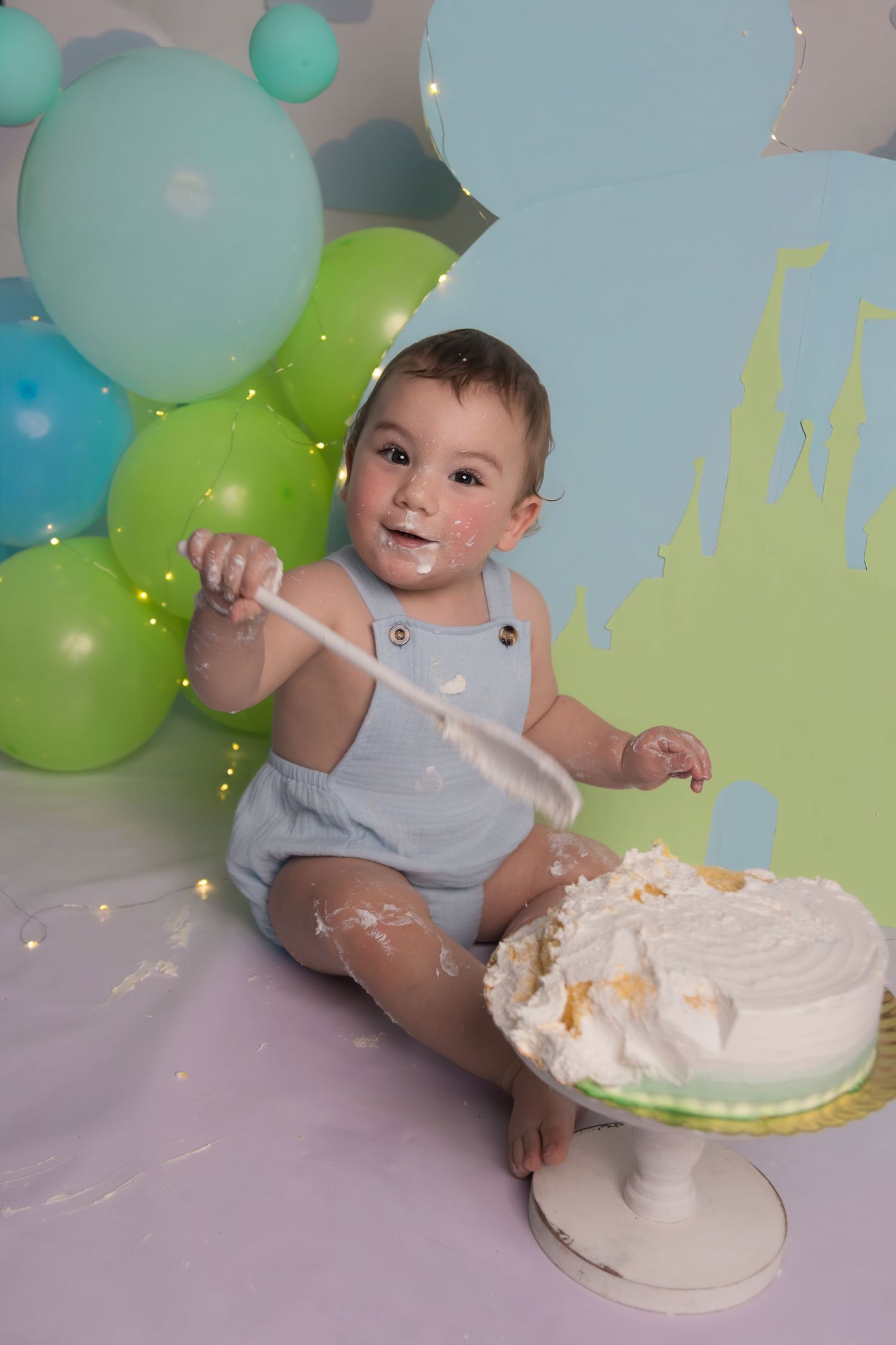 Baby in light blue romper enjoying cake smash photo session with balloons and decorative backdrop.