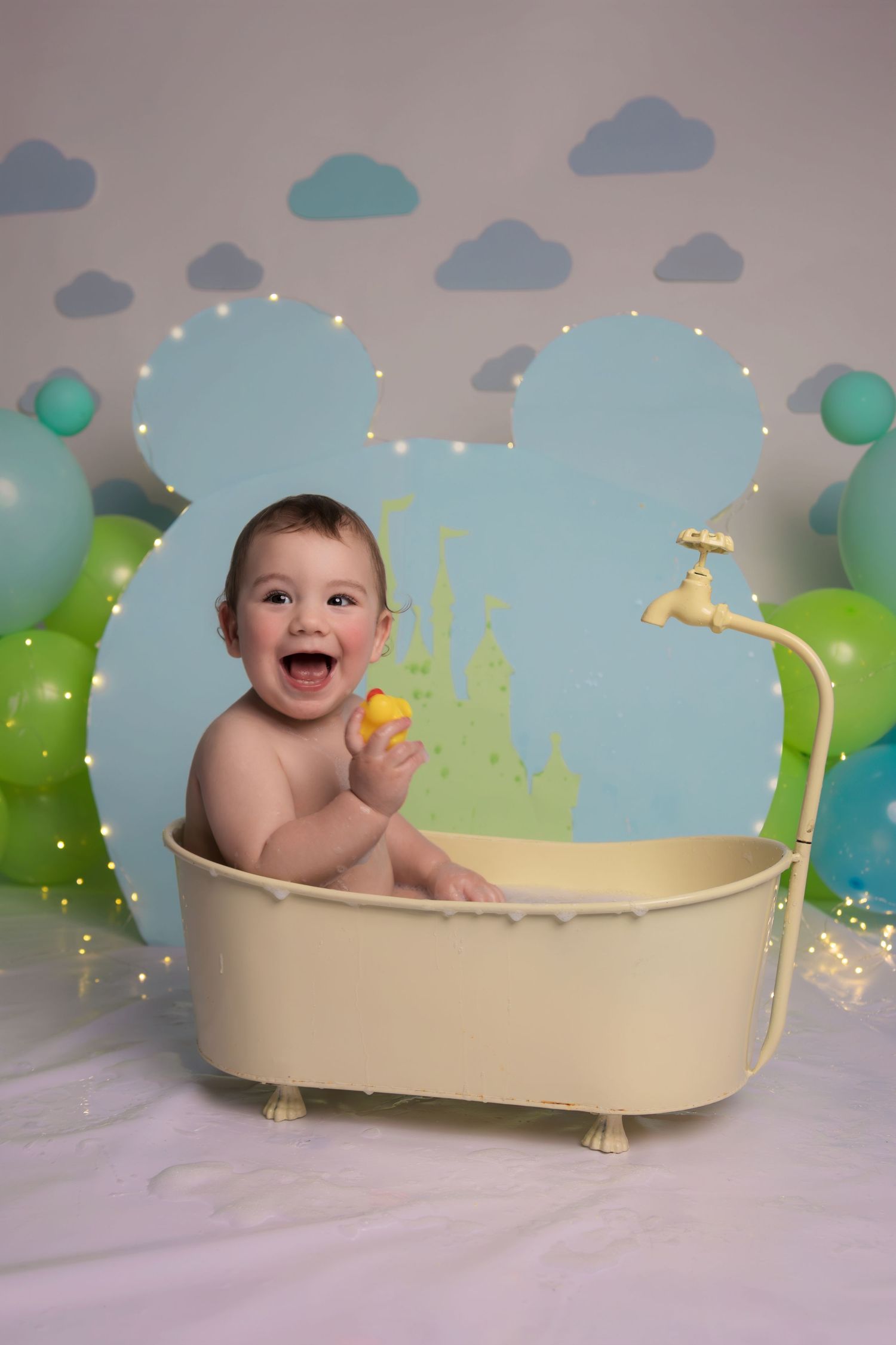 Happy baby sitting in miniature bathtub with rubber duck against blue backdrop with clouds and twinkle lights.