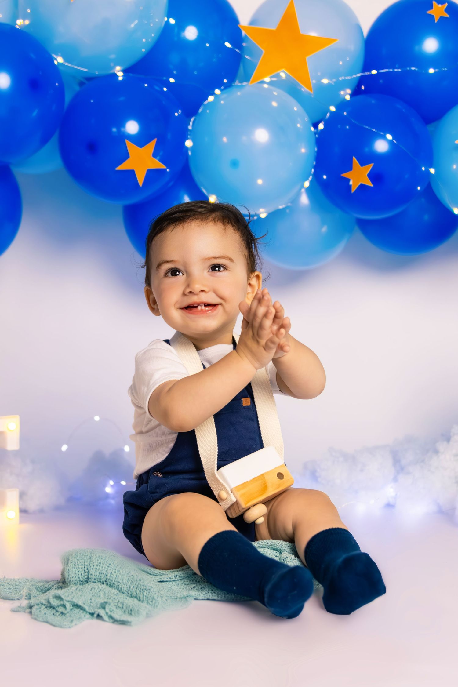 Baby in navy outfit clapping hands against blue balloon backdrop with gold stars.