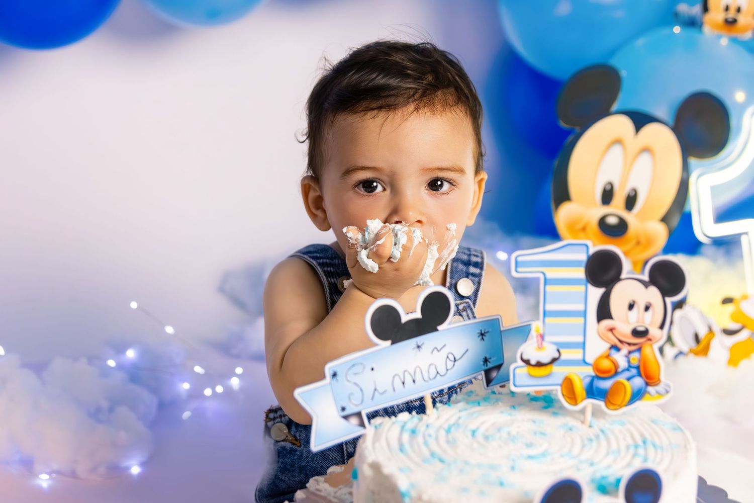 Baby enjoying cake smash with Mickey Mouse themed decorations and blue balloons.