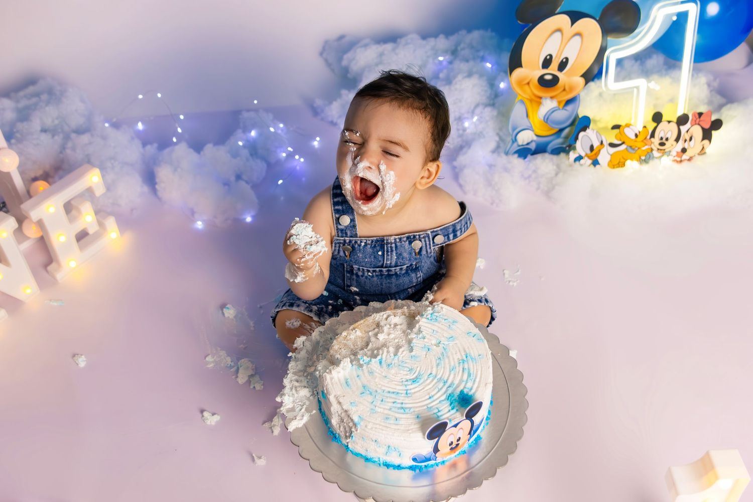 Joyful cake smash session with baby covered in frosting against blue balloon backdrop.