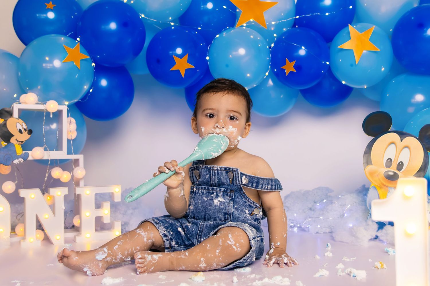 Baby in denim overalls enjoying sweet treats at Mickey Mouse themed photoshoot.