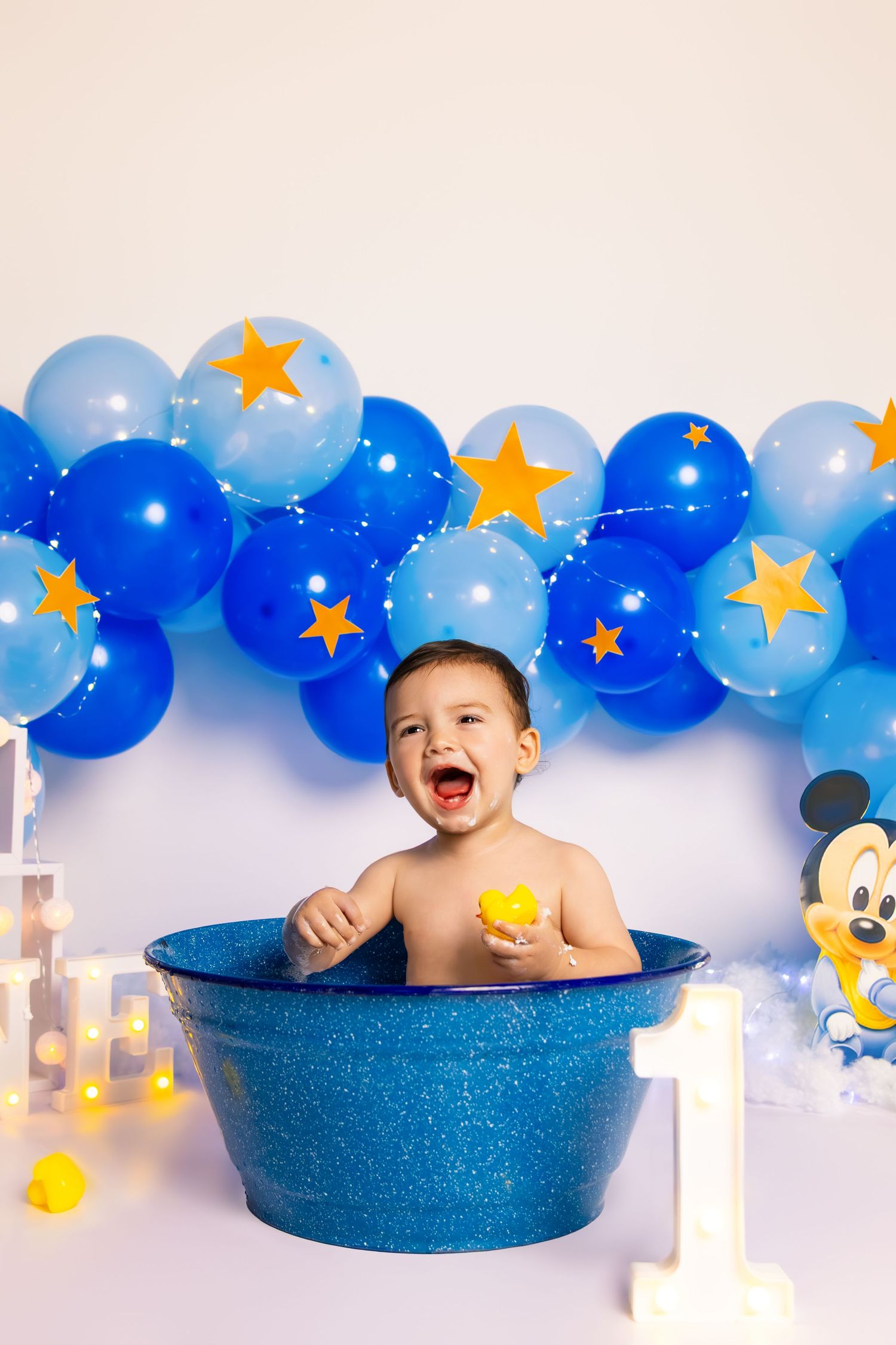 Baby sitting in blue tub with rubber ducks during Mickey Mouse themed first birthday shoot.