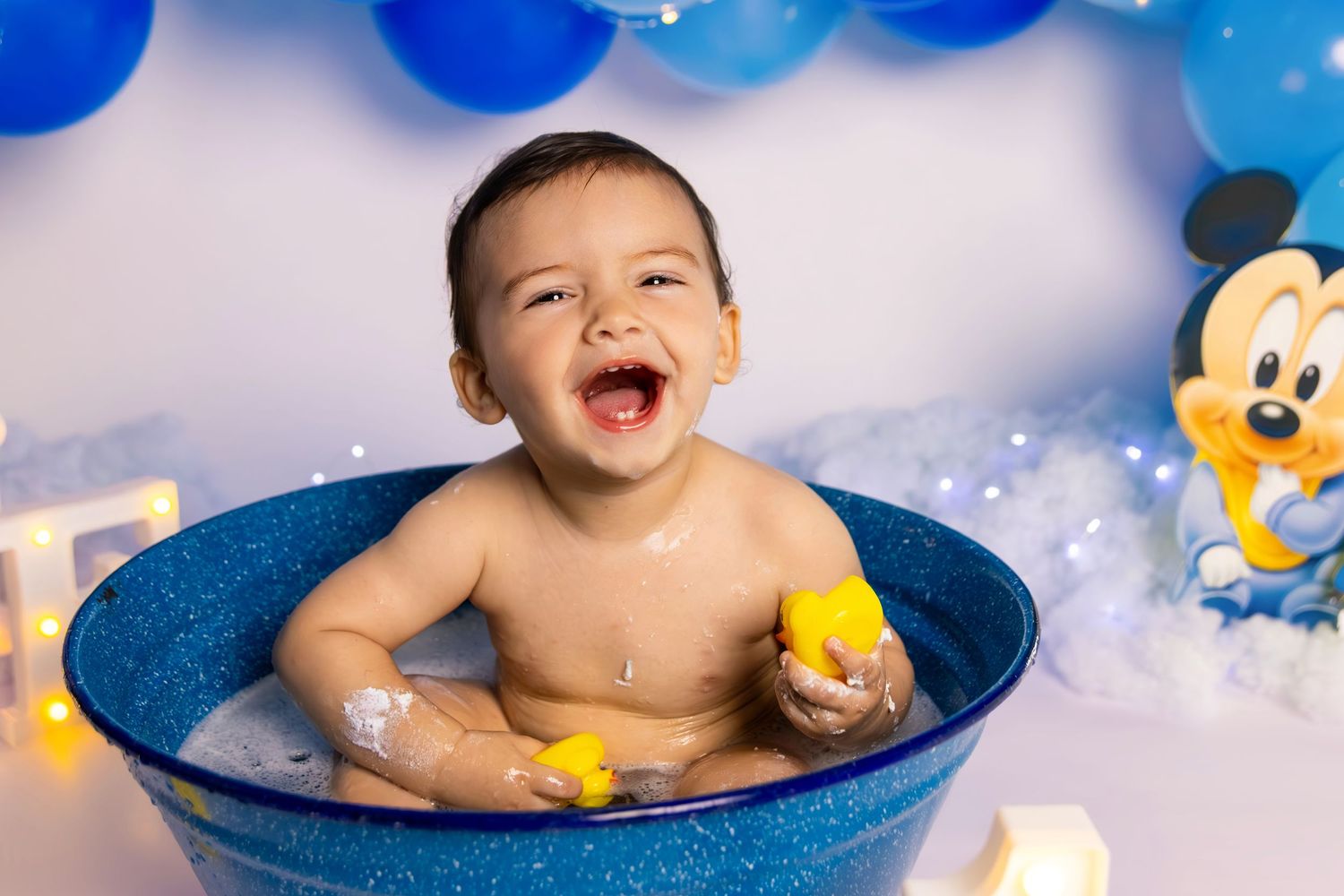 Happy baby splashing in blue tub with rubber ducks during cake smash photoshoot.