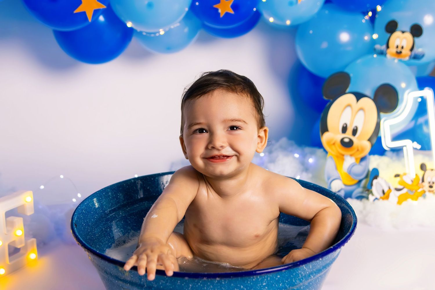 Baby in blue basin with Mickey Mouse themed balloon decorations and stars in the background.