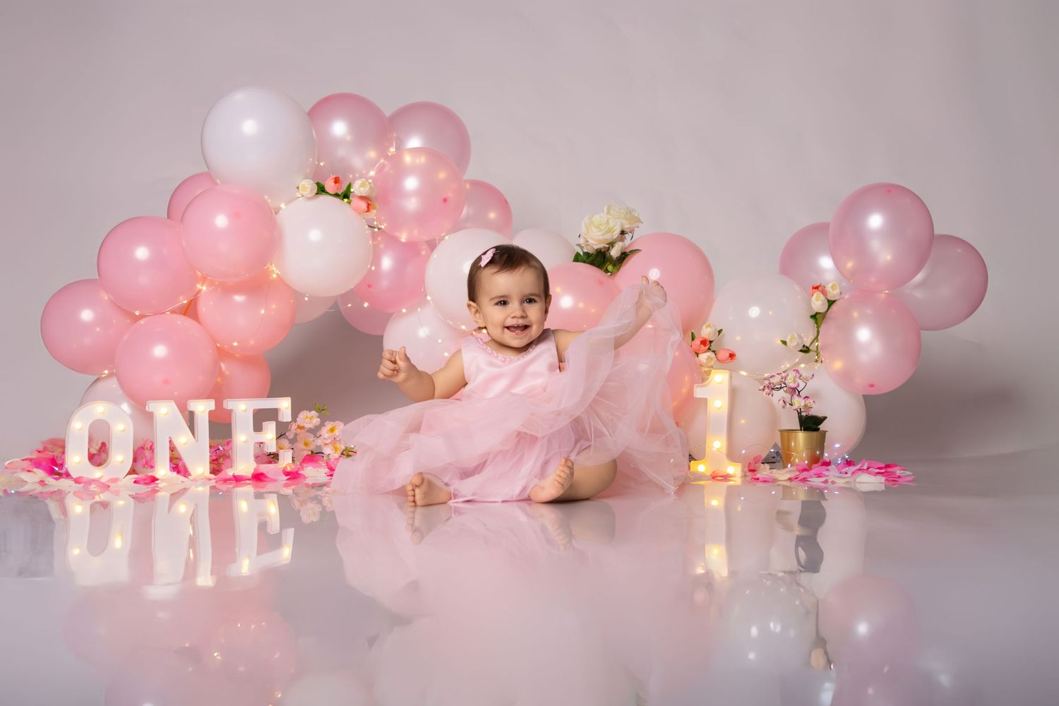 Pink and white balloons with gold accents and string lights create a dreamy first birthday party setup.