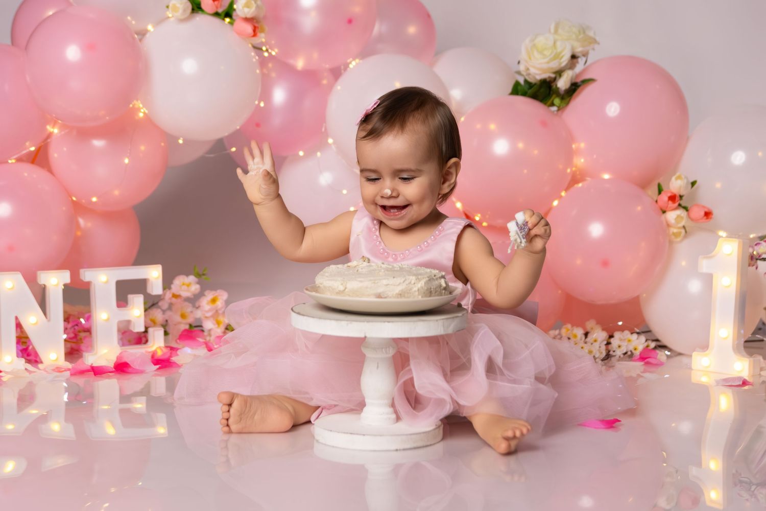 First birthday cake smash setup with pink balloons, string lights and floral decorations.