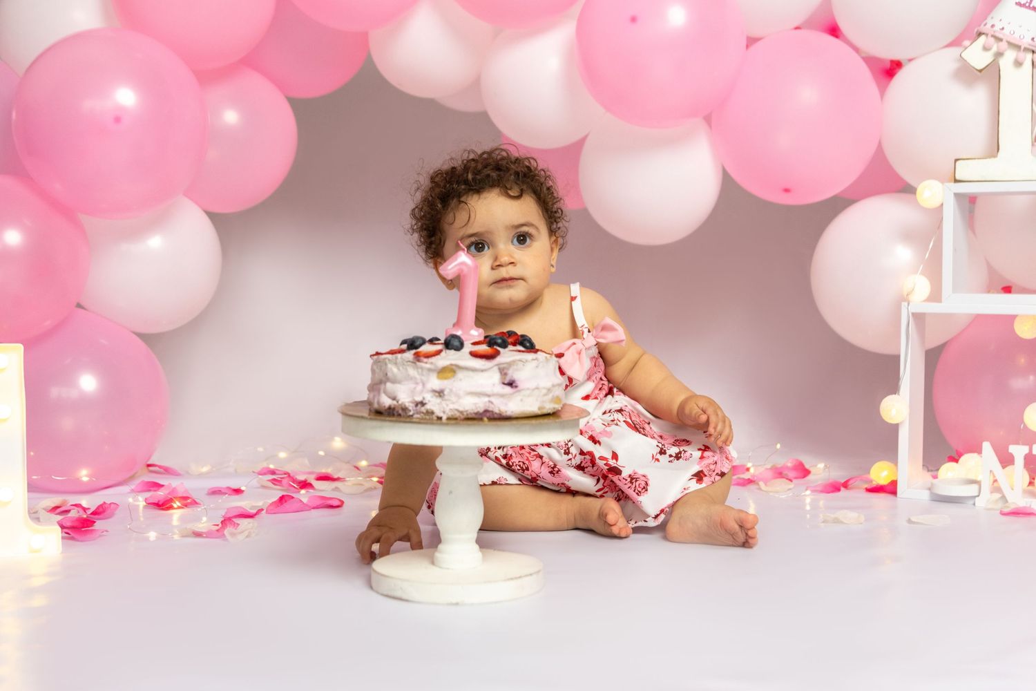 Baby in pink outfit celebrates first birthday with cake and balloon decorations.