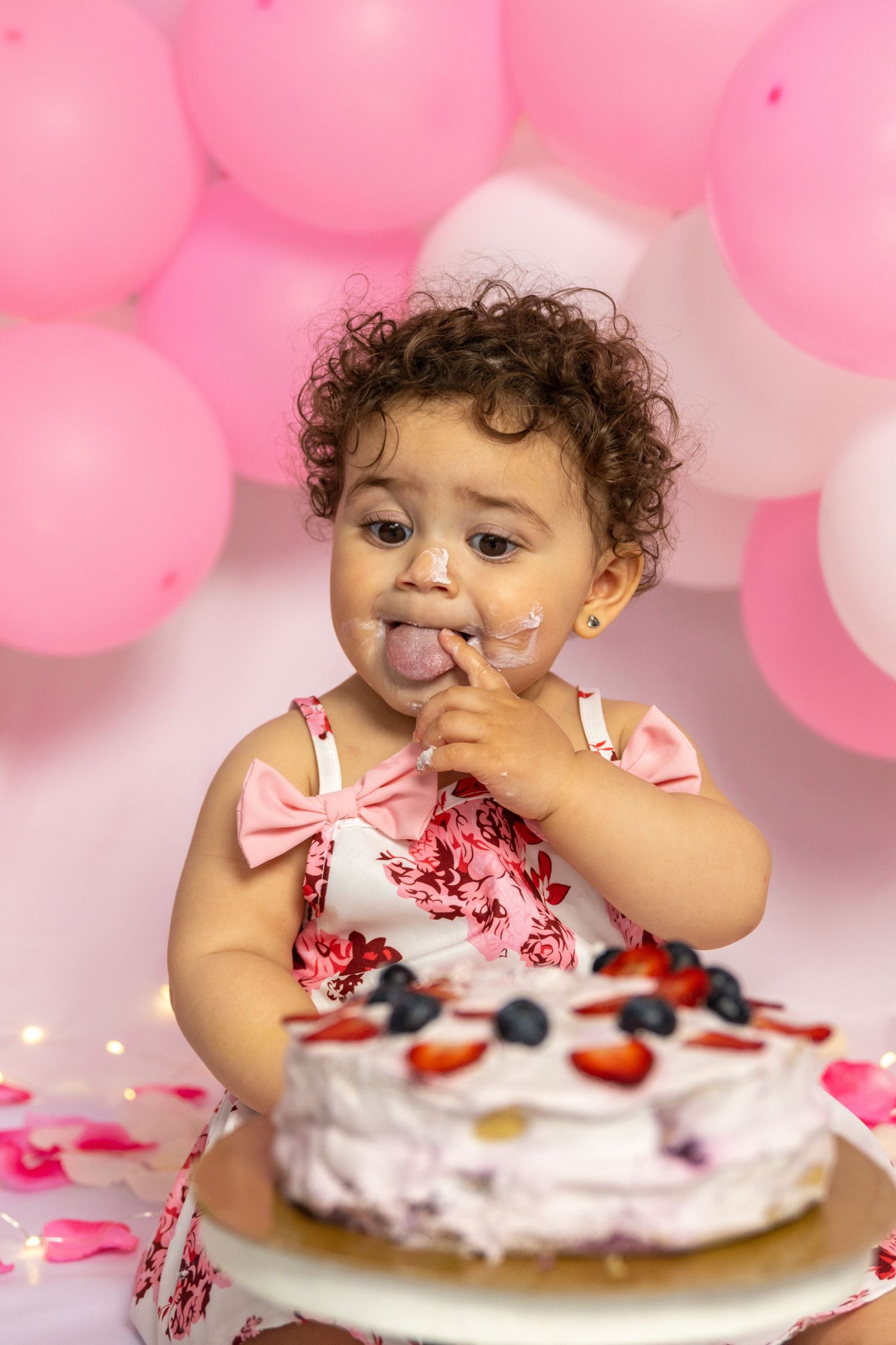 A baby in pink dress enjoying cake with strawberries and blueberries on top.