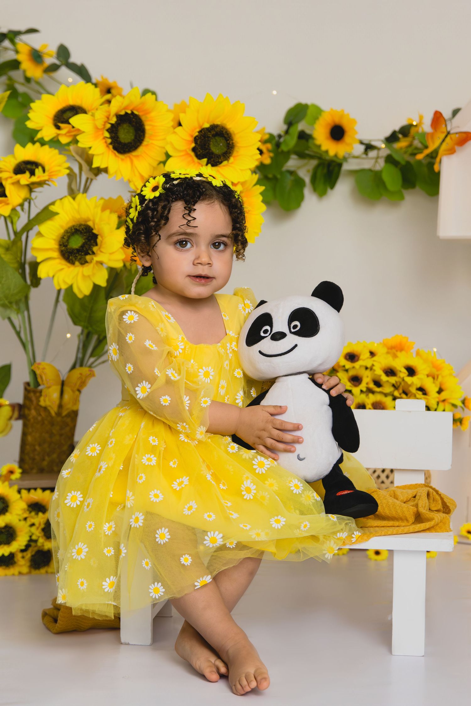 A toddler girl in a yellow daisy dress holds a panda stuffed animal, seated among bright sunflowers.