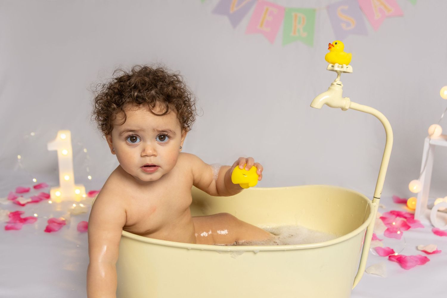 A baby bath photoshoot with rubber ducks and pink flower petals scattered around.