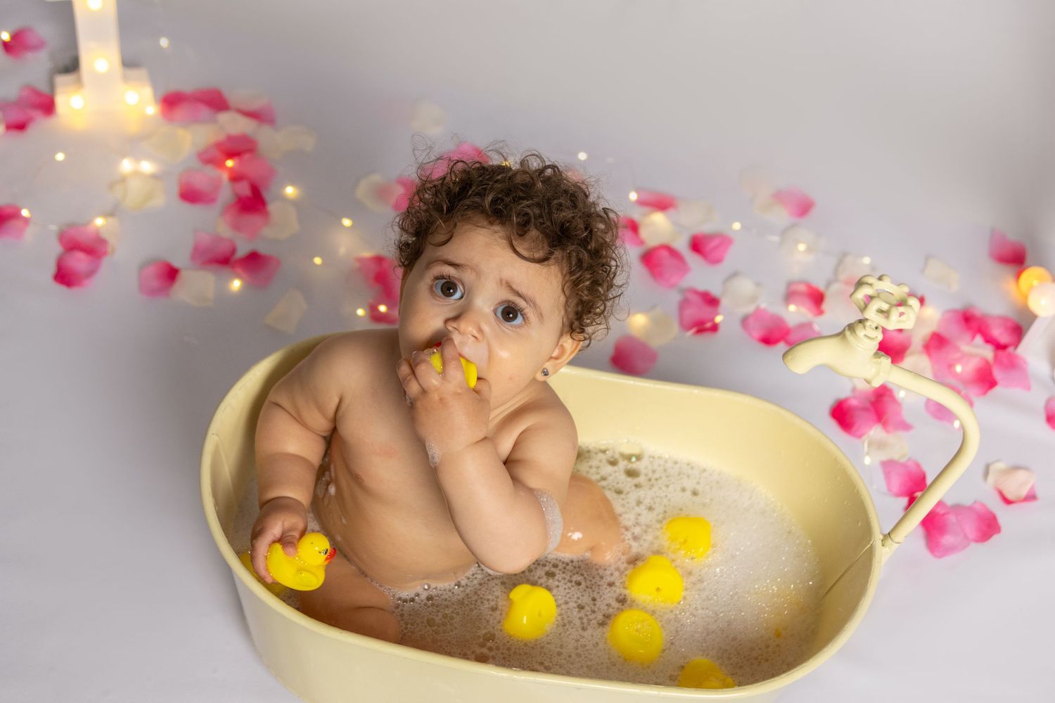 A baby splashing in a cream-colored tub with yellow rubber ducks and rose petals.