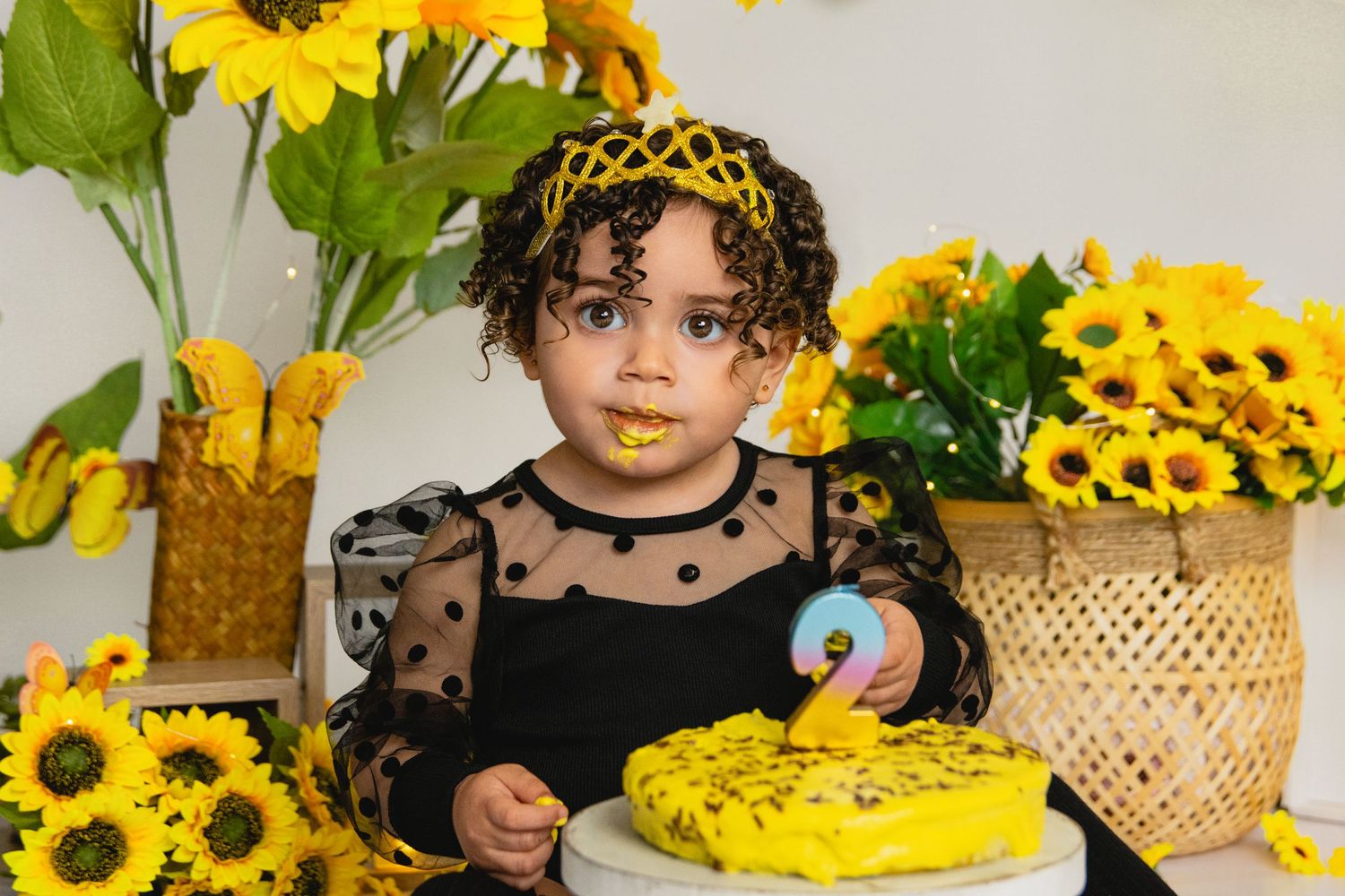 Toddler in black dress with yellow crown celebrates 2nd birthday with sunflower-themed cake smash.