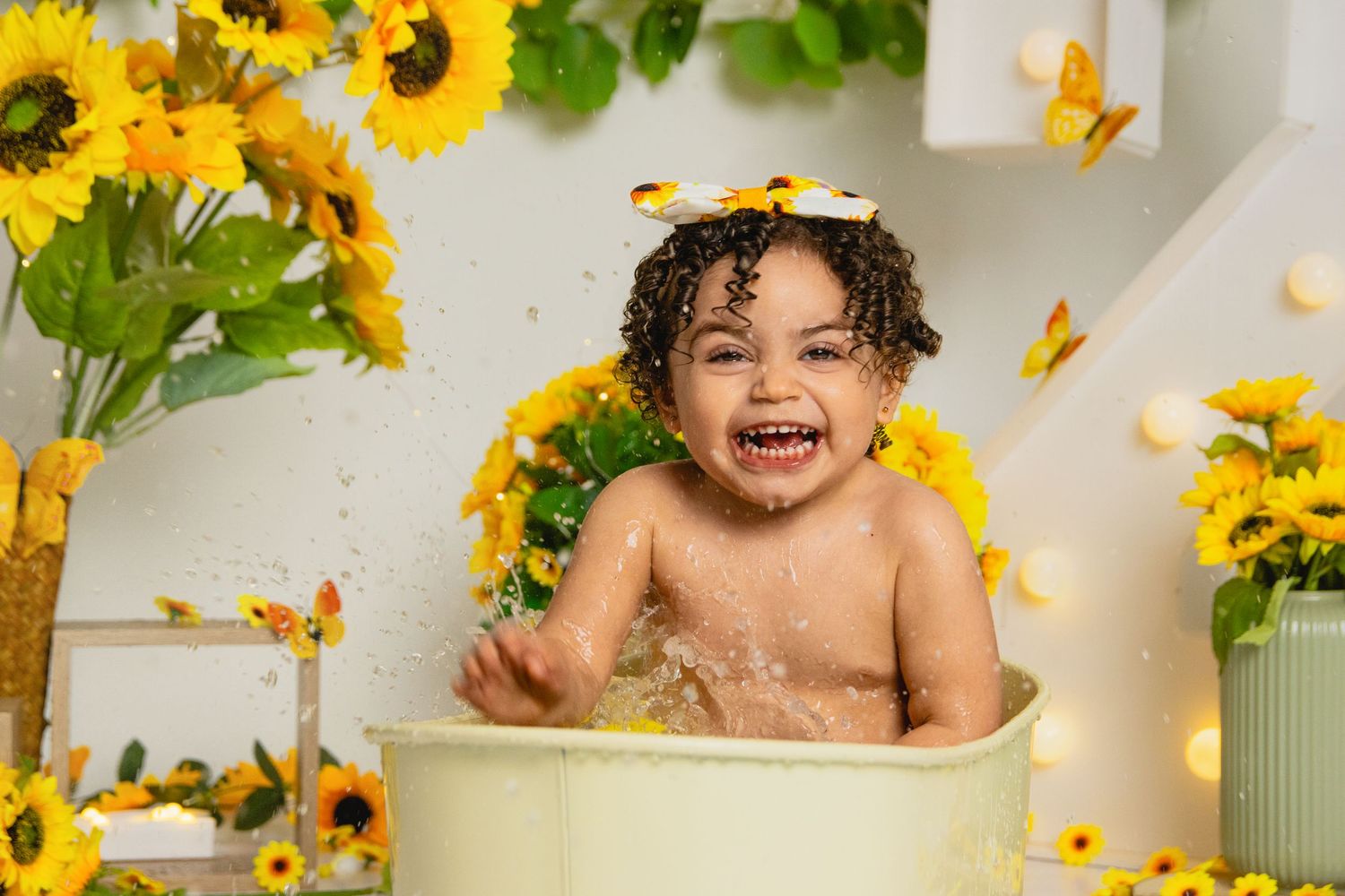 Joyful toddler splashing in a green tub surrounded by sunflowers and butterflies in a bright studio photo.