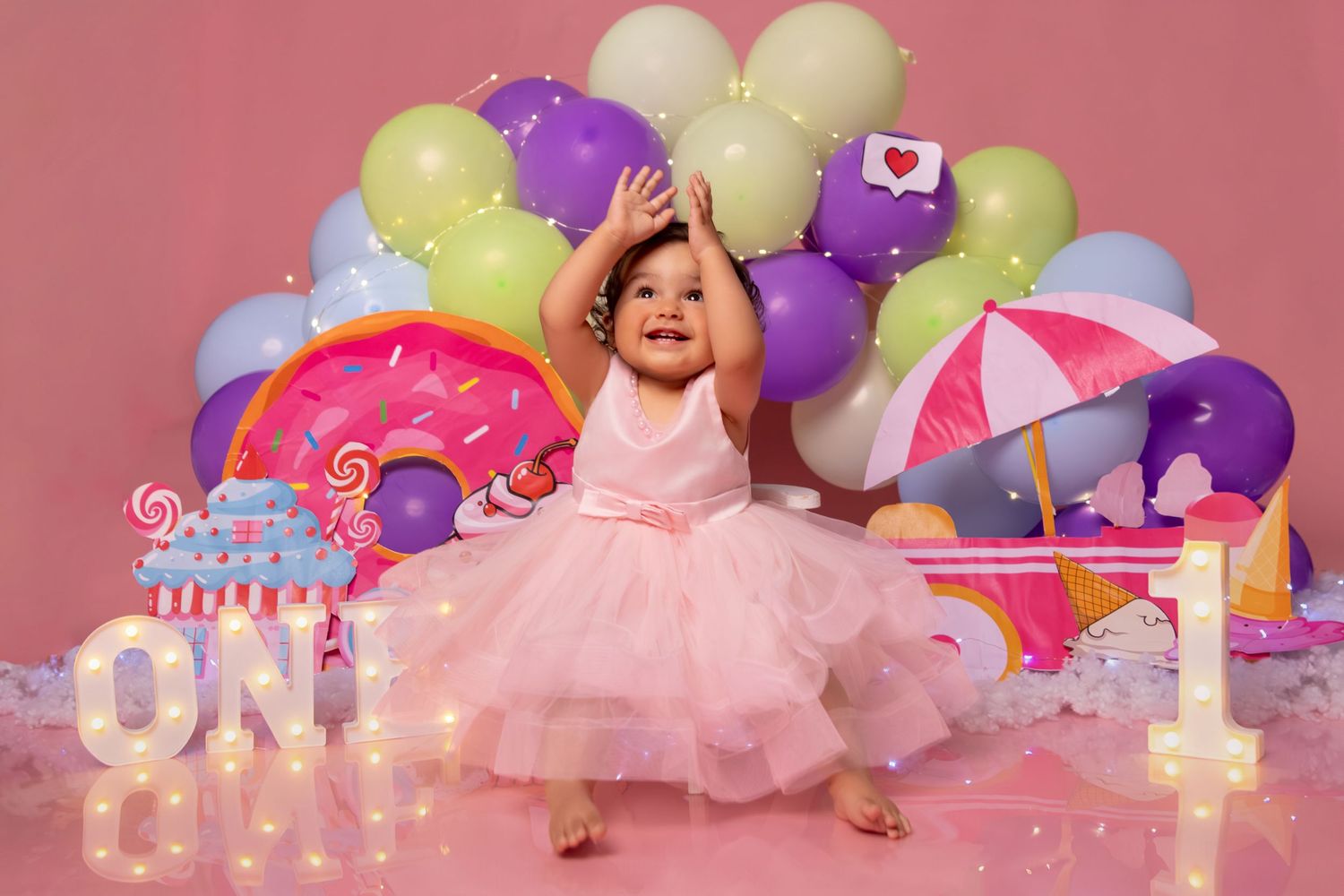 Festive first birthday setup with balloons, cake decorations and pink backdrop.