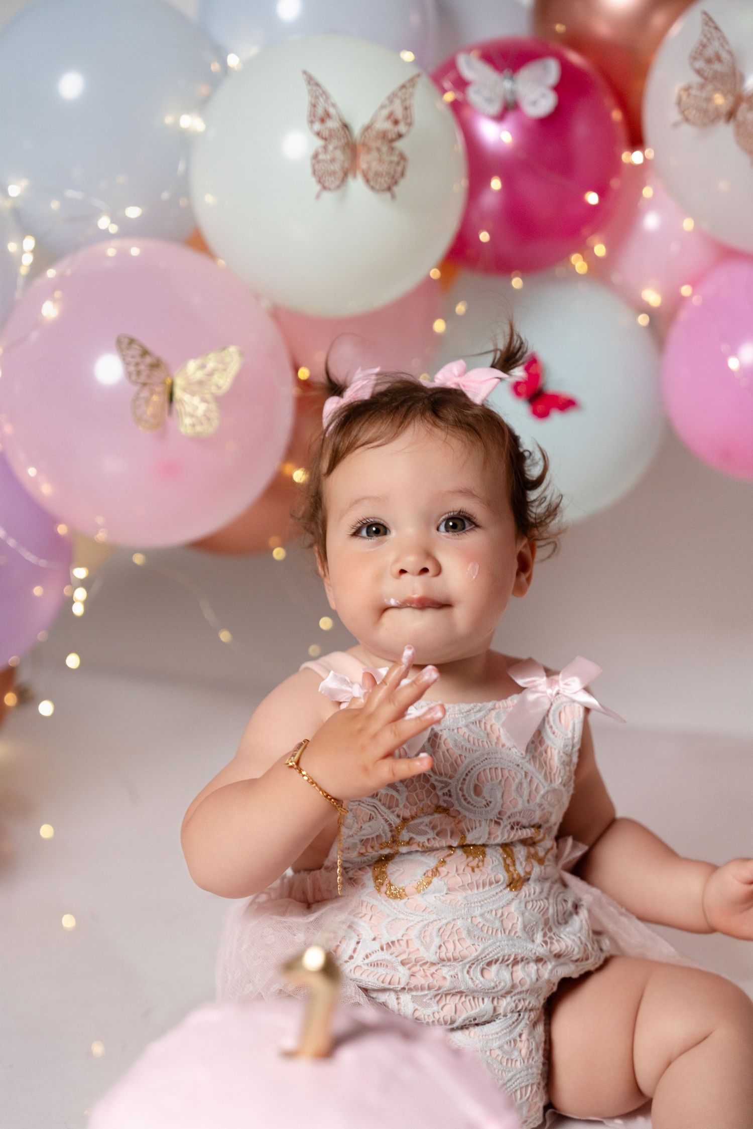 Adorable baby girl in pink lace dress celebrating first birthday with colorful butterfly balloons.
