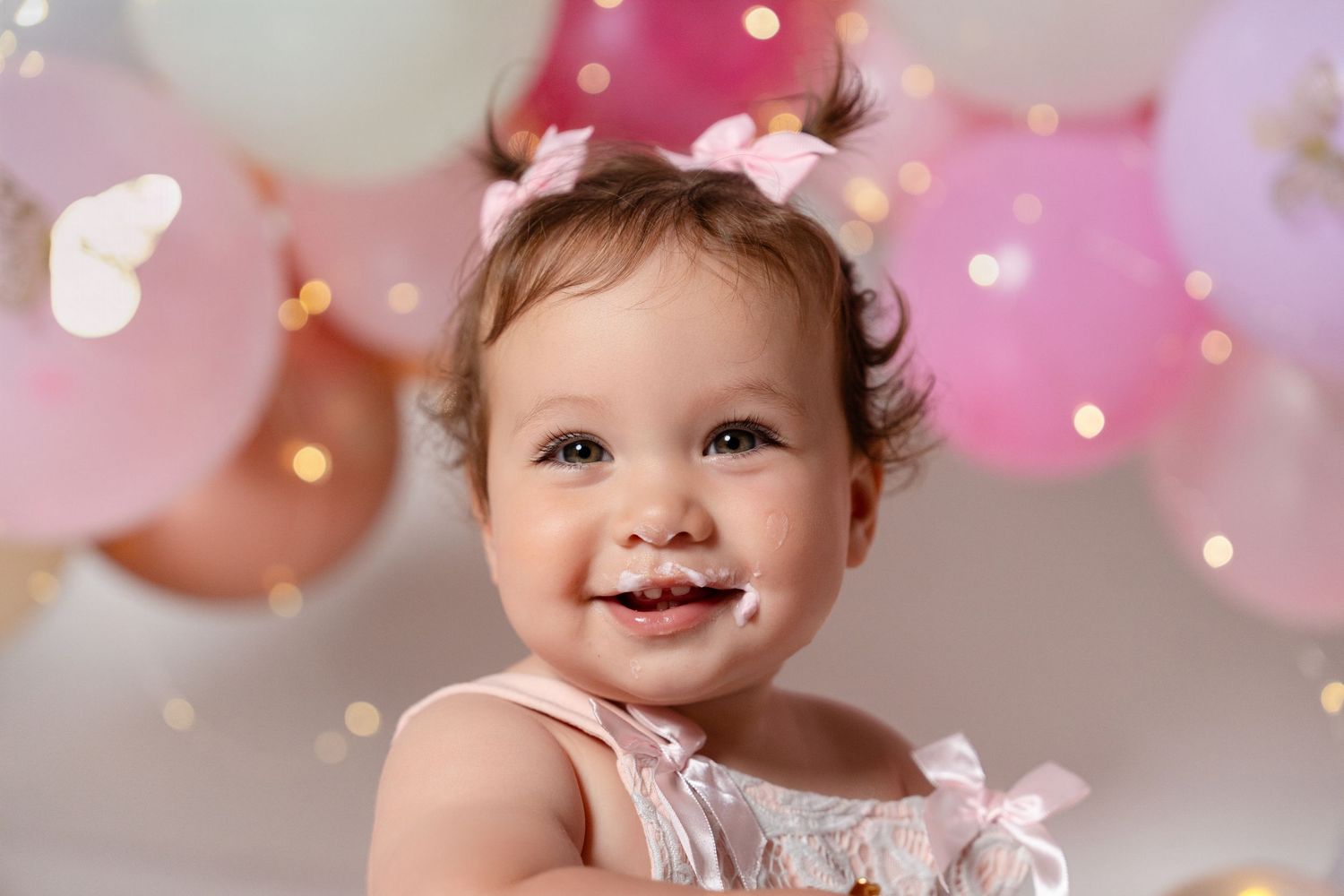 Smiling baby girl with pink bow celebrating first birthday surrounded by pink and gold balloons.