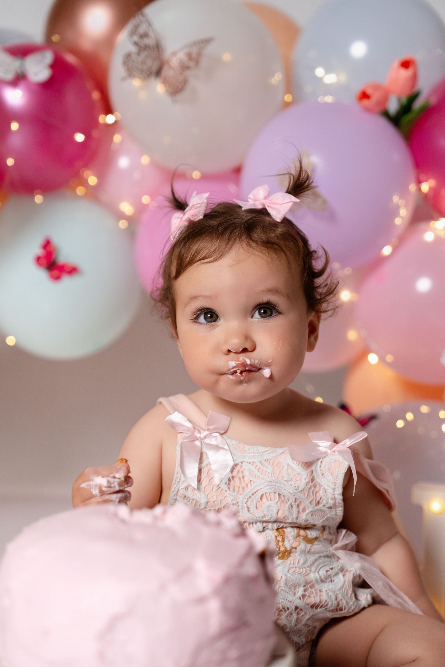 Baby girl in lace romper eating first birthday cake surrounded by pink balloons and butterfly decorations.