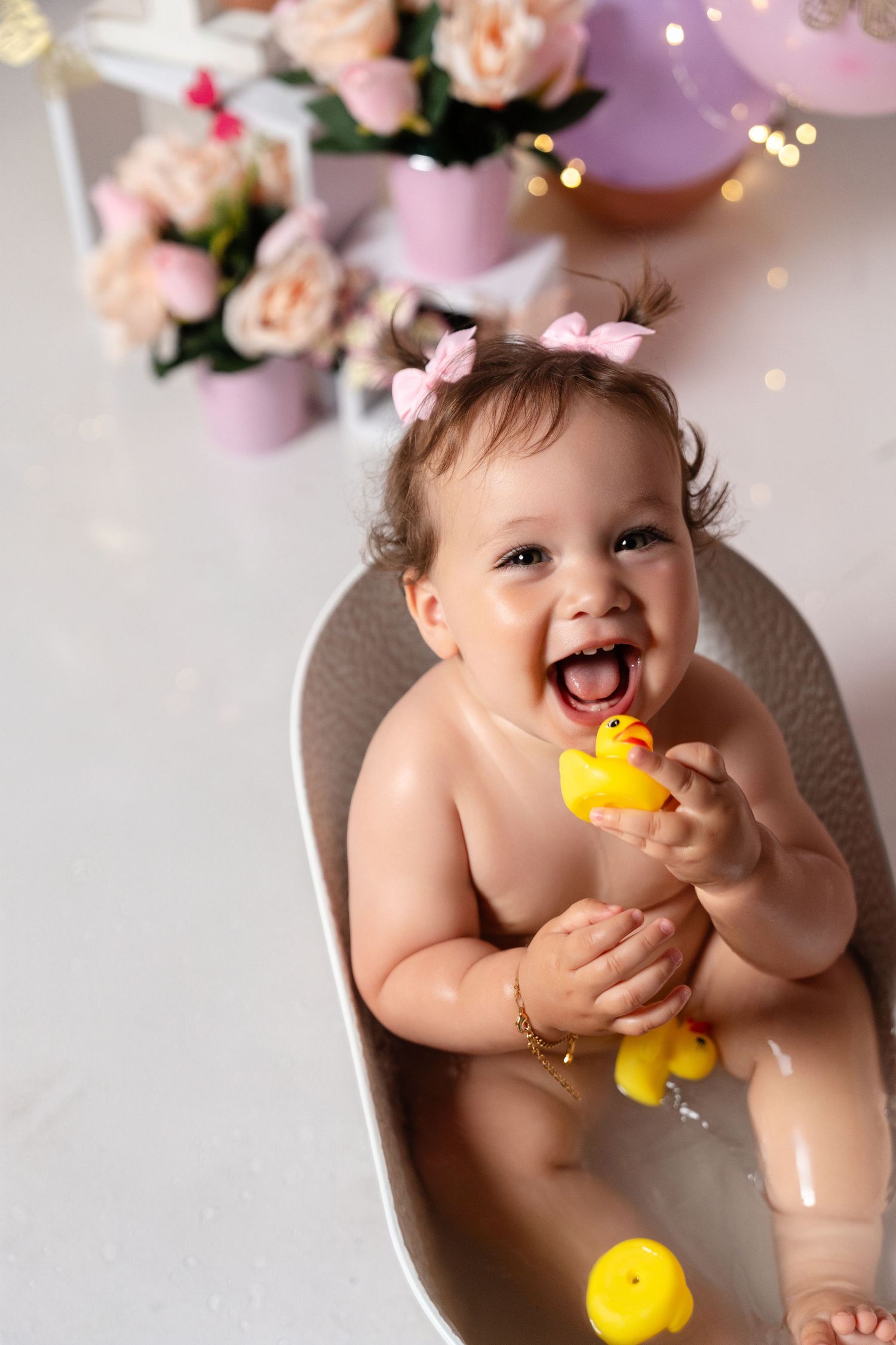 Happy baby girl laughing in small bathtub holding yellow rubber ducks, with pink bows in hair and floral decorations behind.