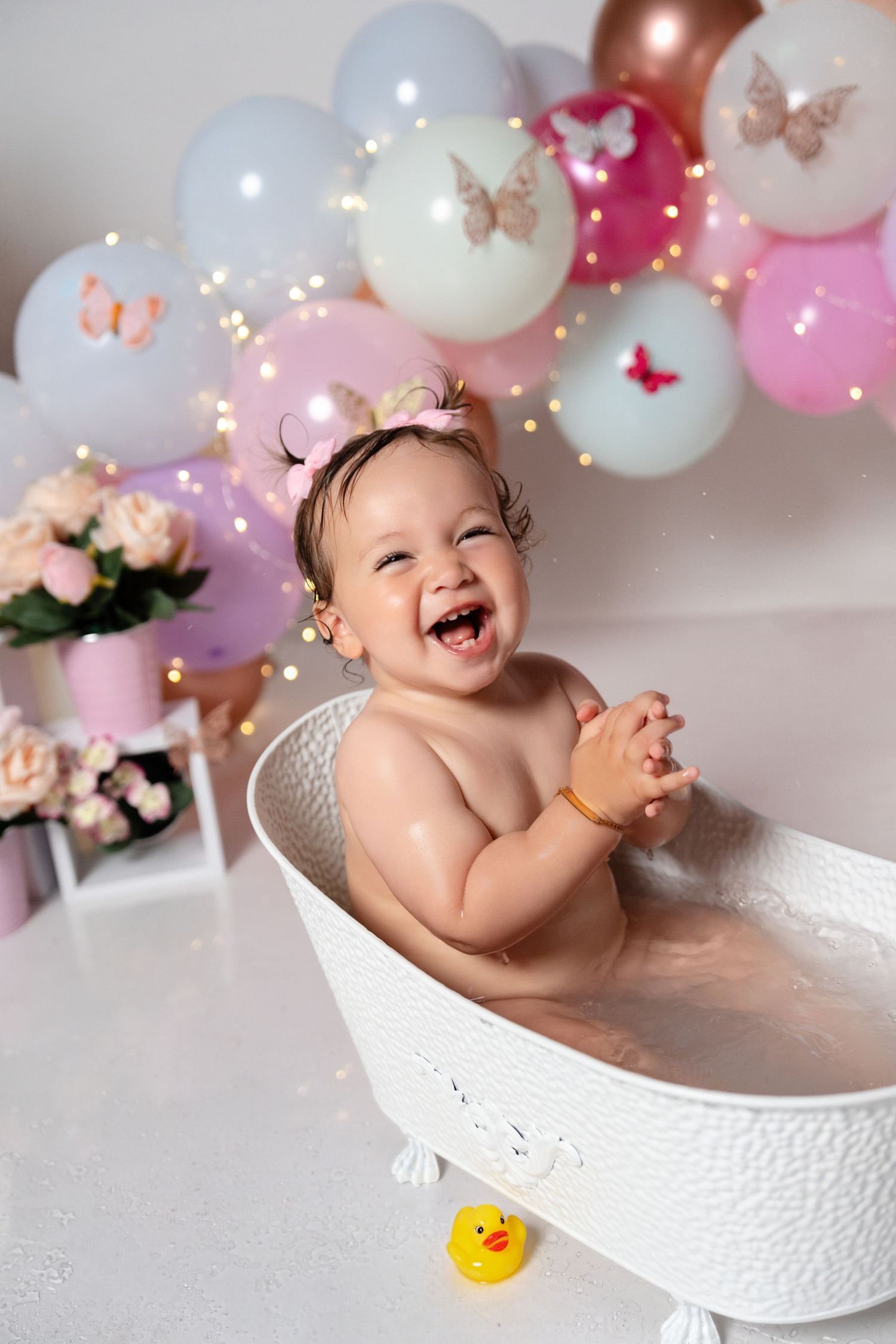 Happy baby girl laughing in a white bathtub surrounded by colorful balloons and fairy lights at a first birthday celebration.