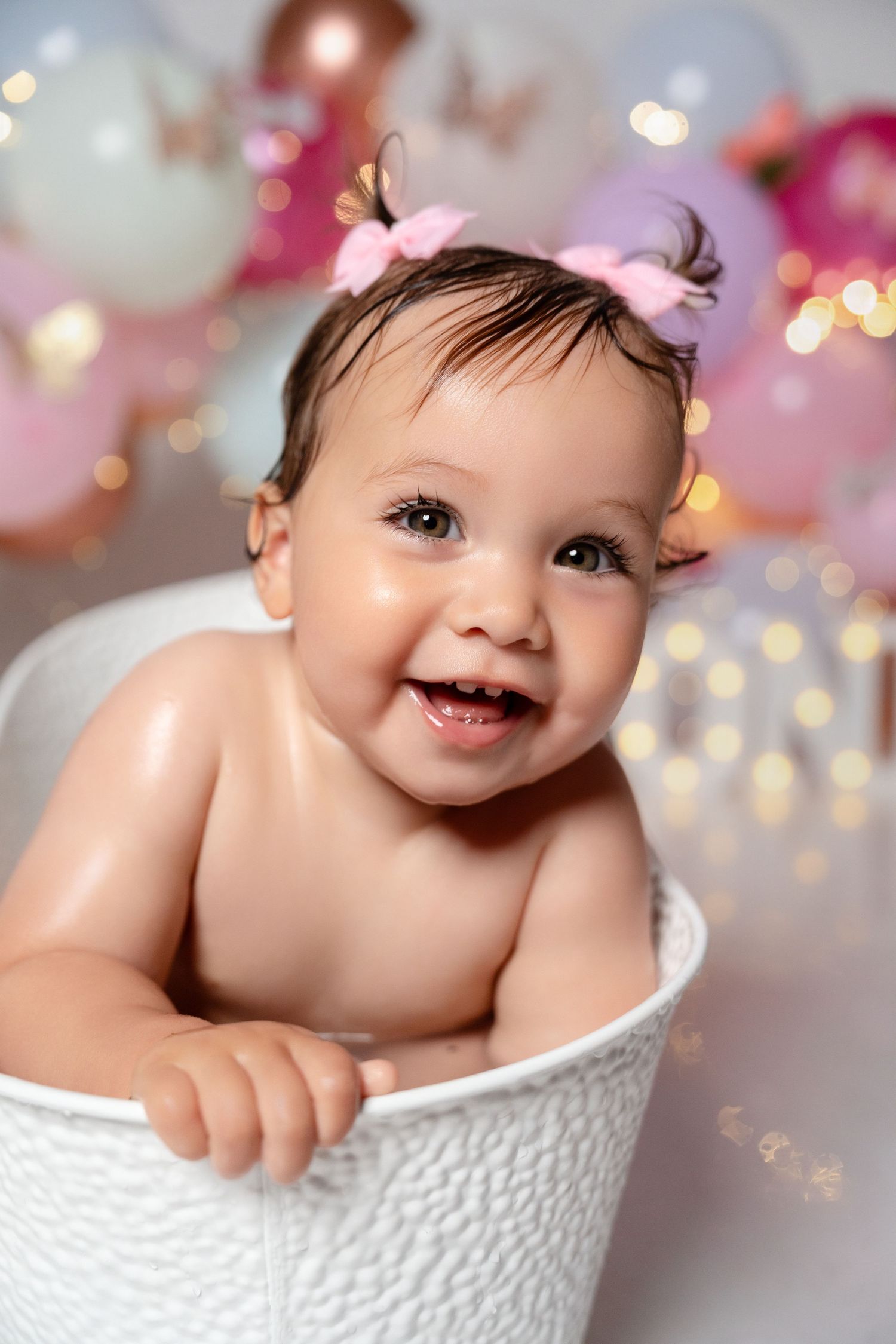 Smiling baby girl with pink bow headband sitting in white bucket tub with colorful bokeh balloons in background.