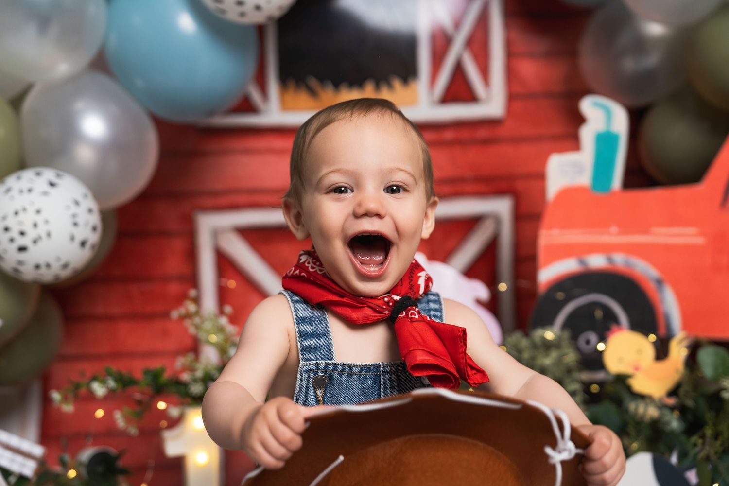 Joyful baby boy in denim overalls and red bandana laughing at first birthday cake smash with farm-themed backdrop.