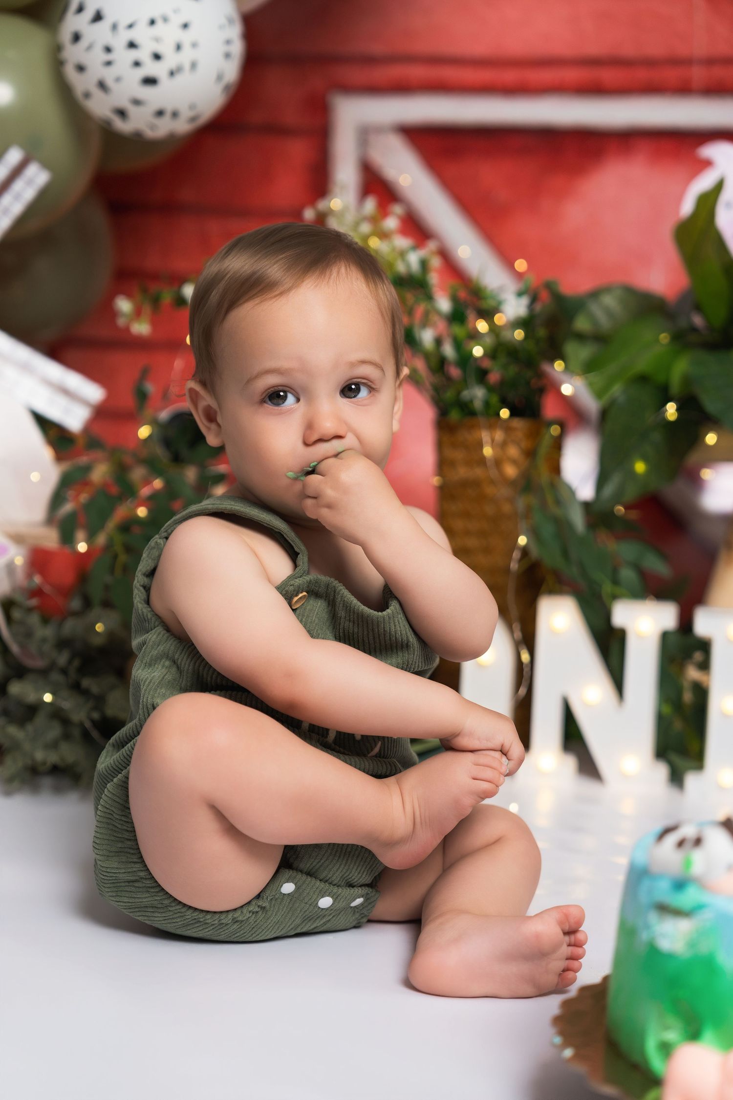 Cute baby in green romper sitting barefoot at a first birthday cake smash with festive green and red decor.