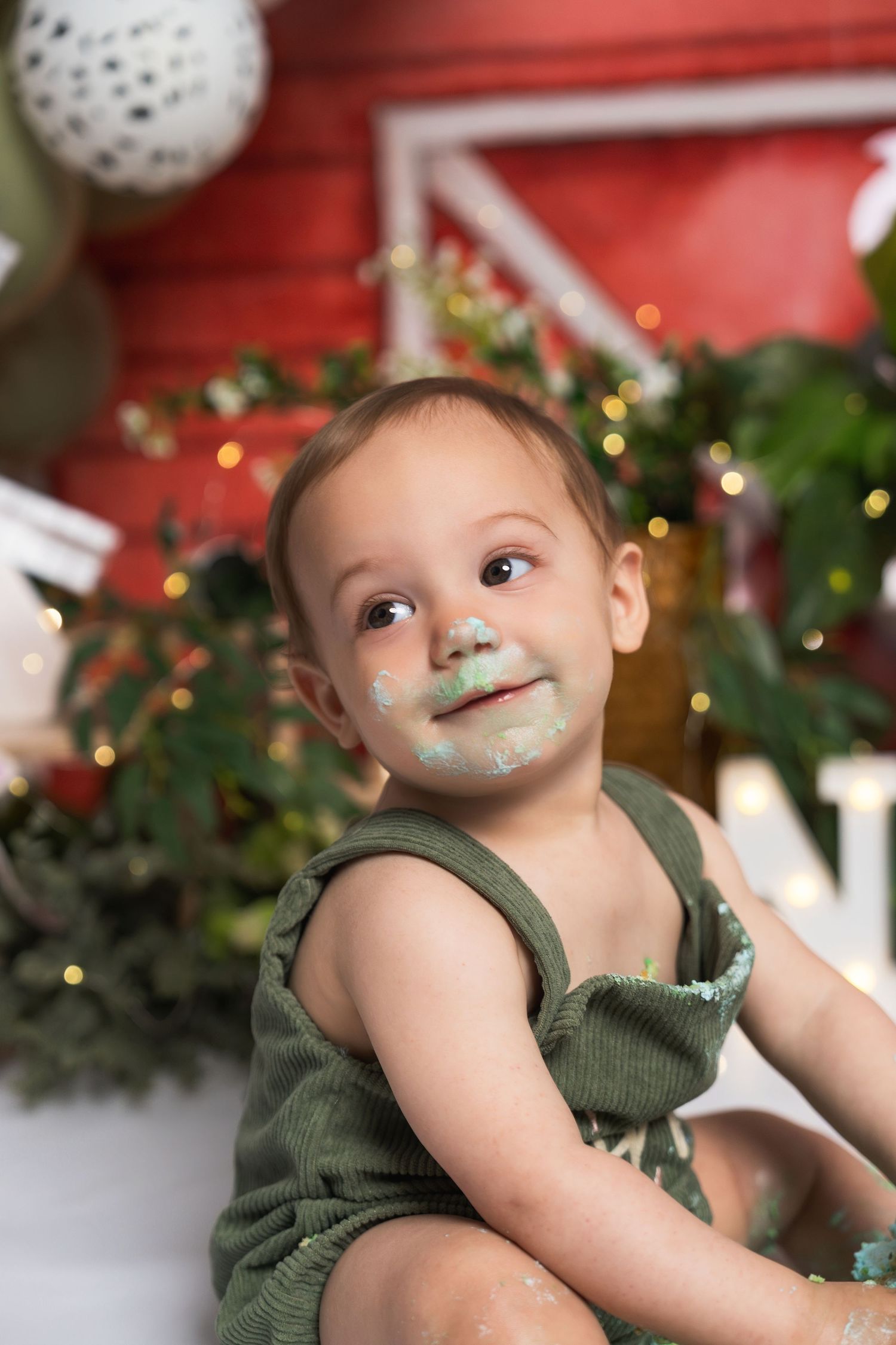 Adorable baby with cake frosting on face sitting in front of festive Christmas holiday backdrop with green plants.