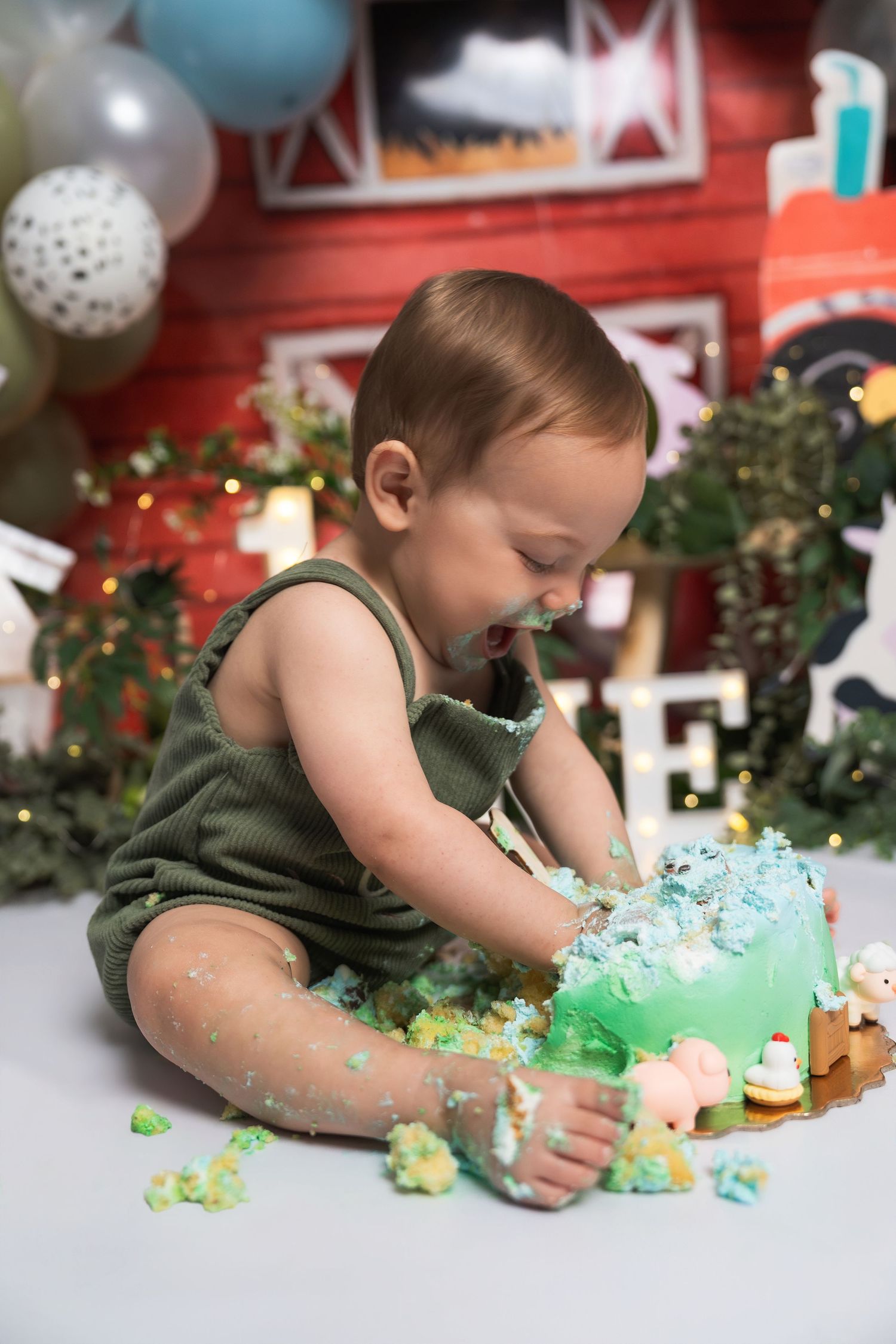 Happy baby in green romper smashing a blue first birthday cake at a farm-themed cake smash photo session.