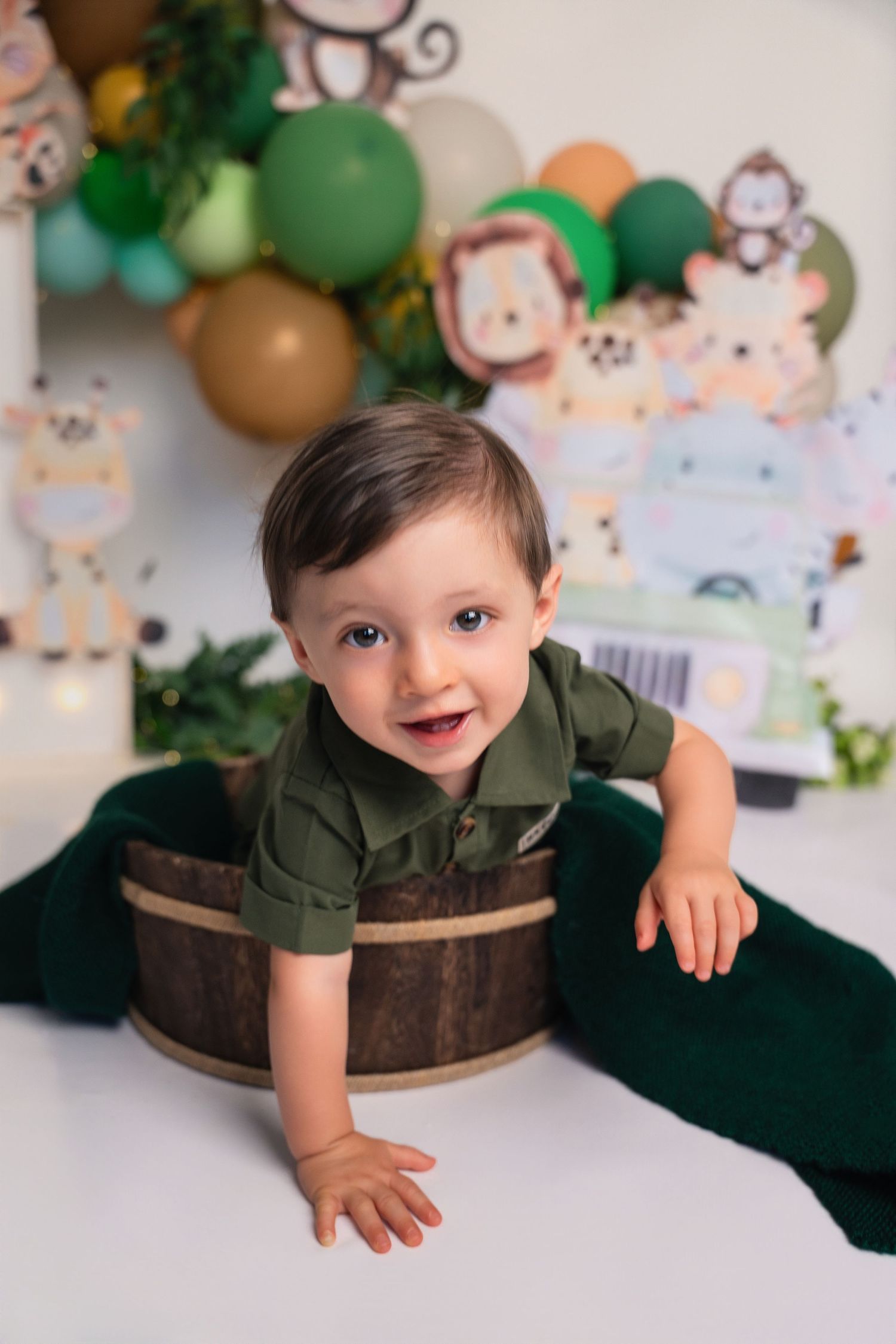 Smiling baby boy in green shirt sitting in wooden barrel with safari balloon backdrop for first birthday.