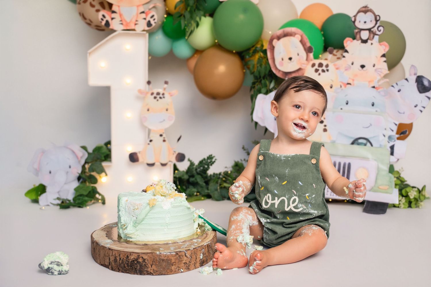 Baby's first birthday cake smash with safari jungle theme, green balloons, and animal decorations on white background.