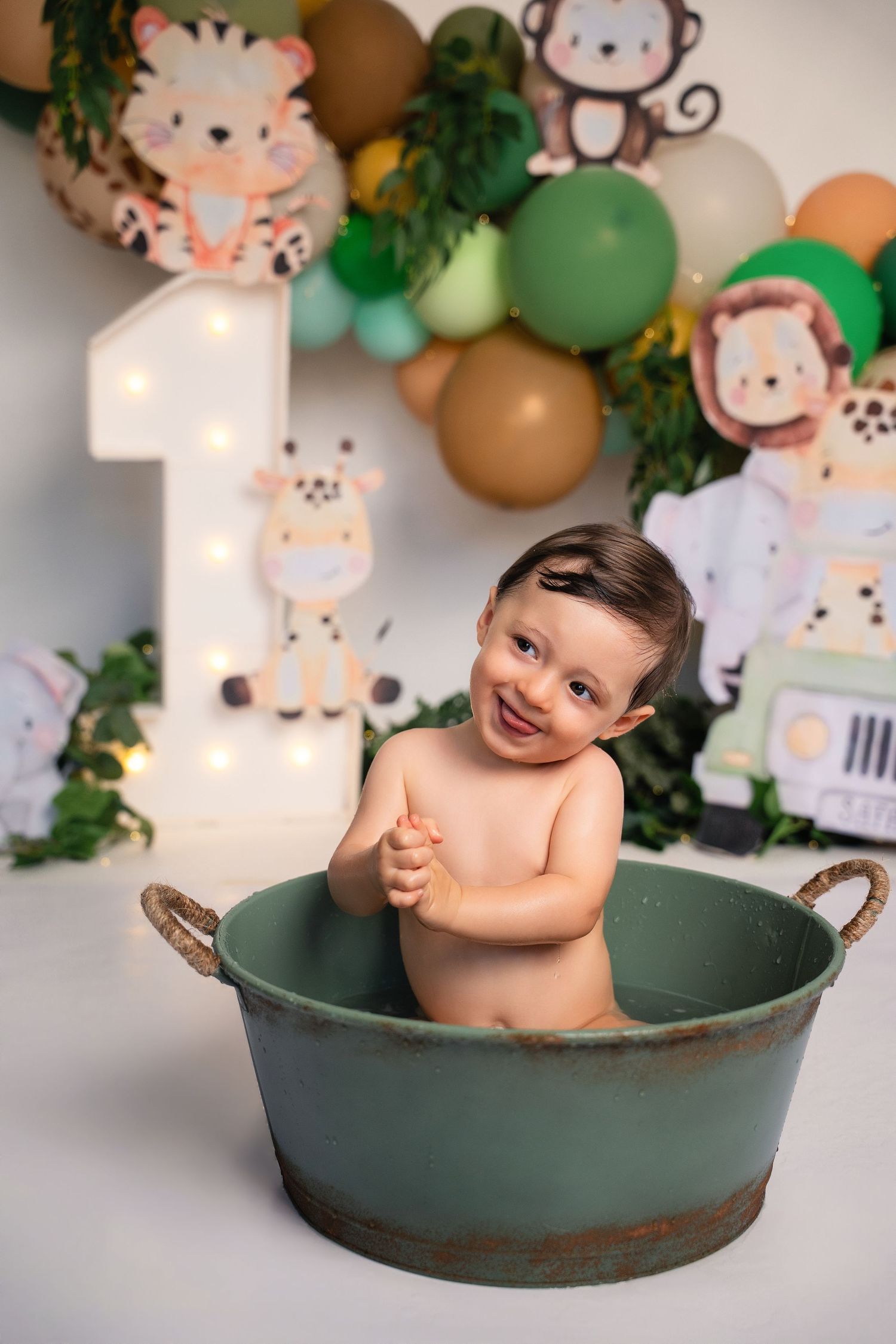 Smiling baby in green tub at safari-themed first birthday party with balloon garland and animal decorations.