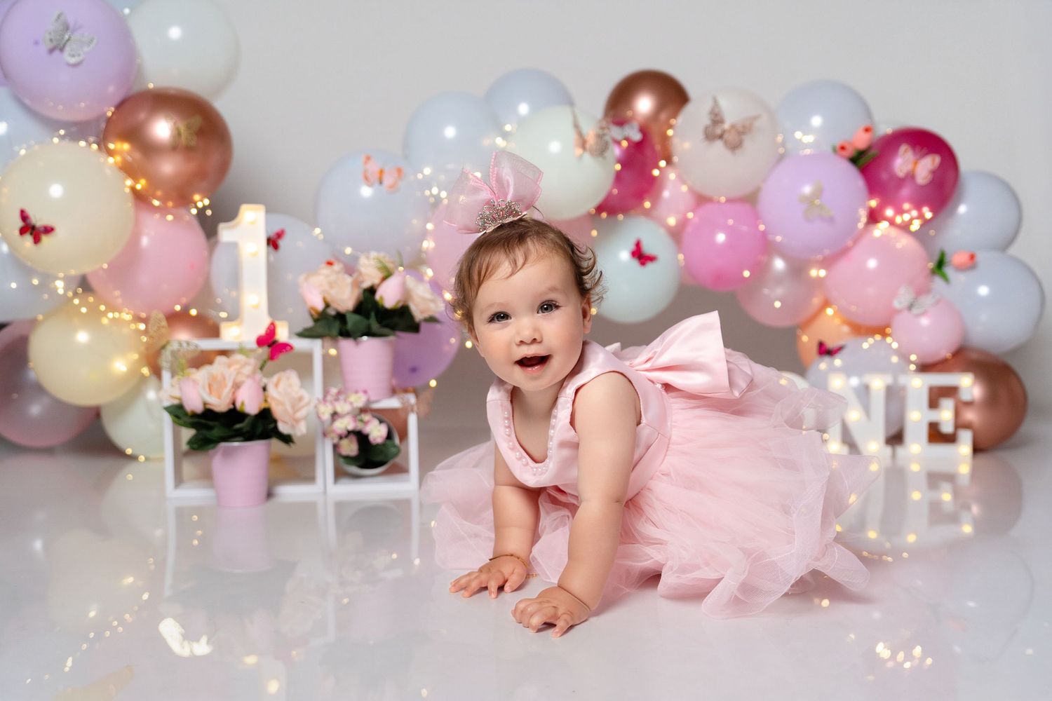 A baby girl in a pink tutu dress crawls on a white floor surrounded by colorful balloons and floral decorations.