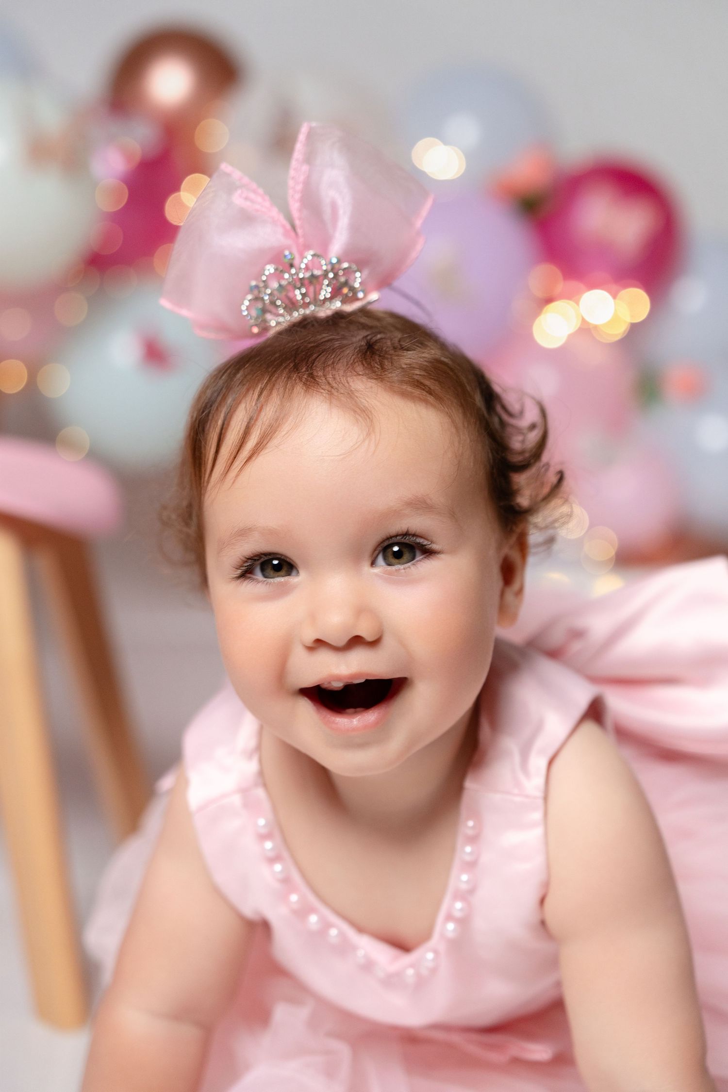 Smiling baby girl in pink dress and tiara crown headband celebrates first birthday with colorful balloons in background.